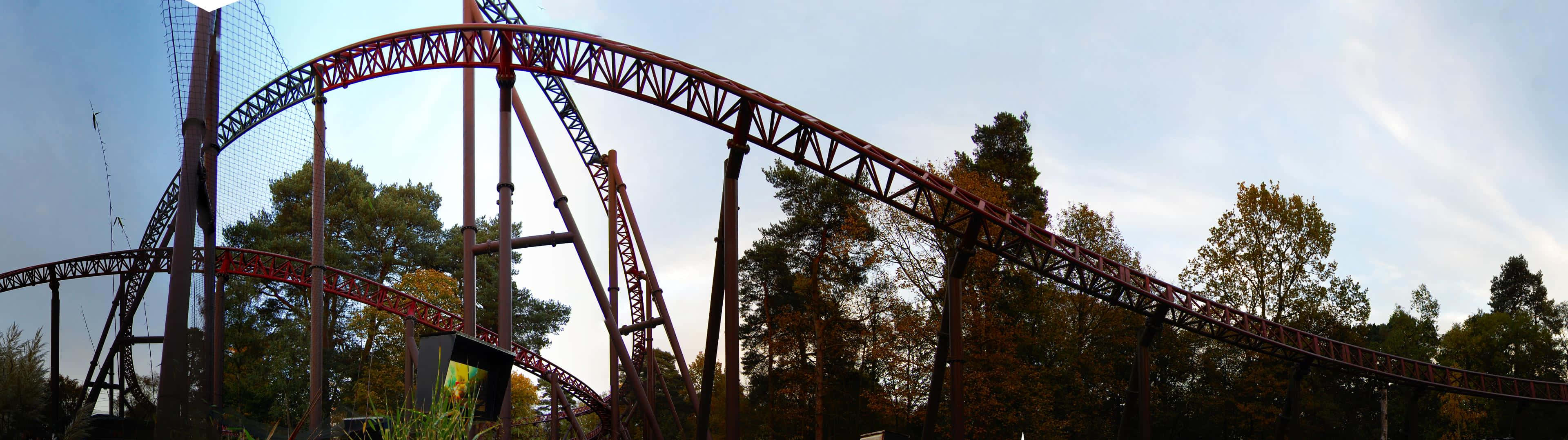 Roller Coaster 桌布 壁紙 — 在淡藍色的天空下，一座高聳的紅色鋼製雲霄飛車在秋樹間蜿蜒穿行。