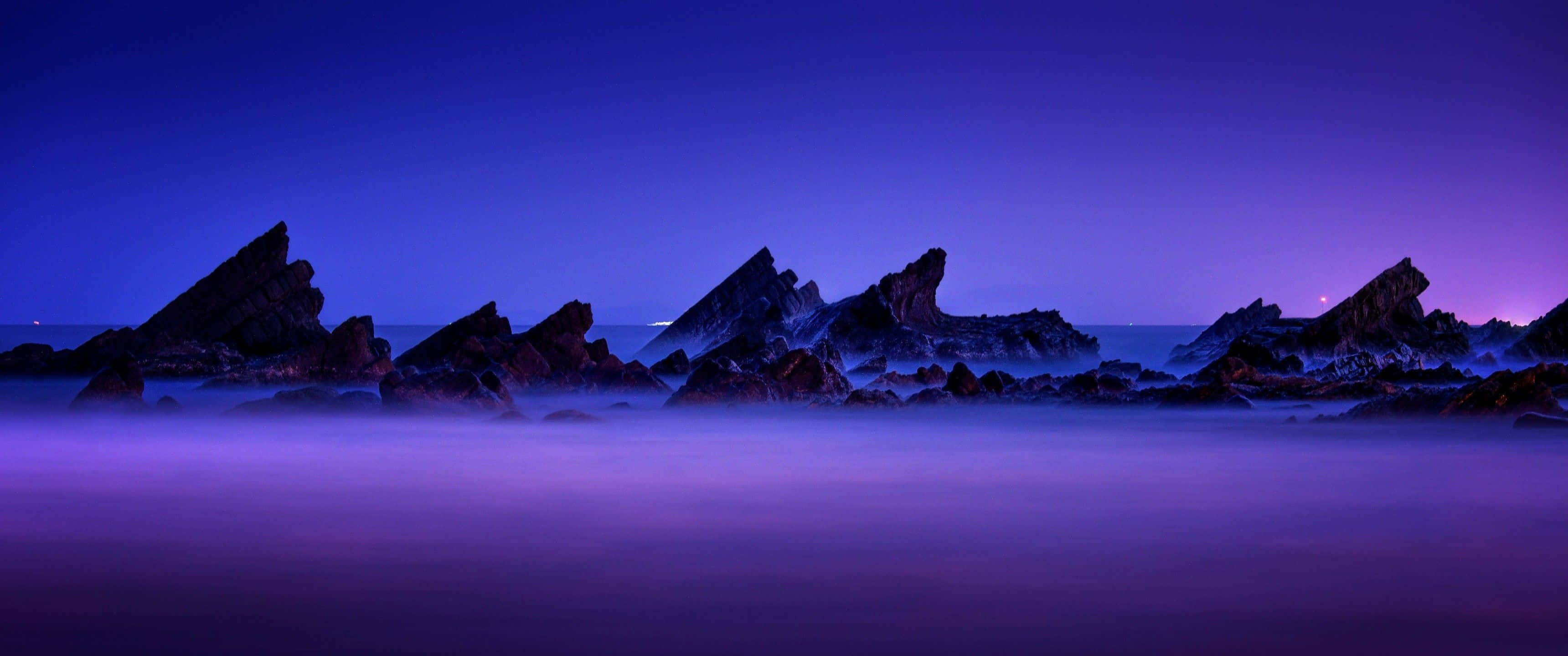 Aesthetic Nature 桌布 壁紙 — 在深沉的暮色天空下，輪廓分明的鋸齒狀岩石從霧氣瀰漫的紫色海洋中升起。