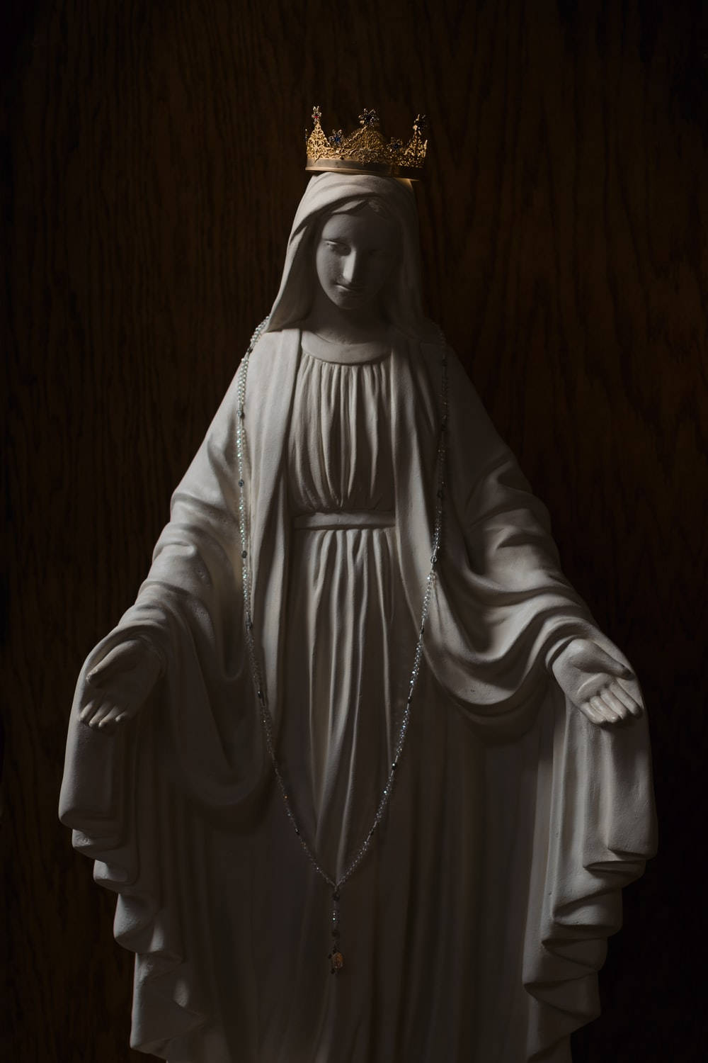 Statue Aesthetic 桌布 壁紙 — 在深色木質背景前，聖母瑪利亞的寧靜白色雕像，戴著金色皇冠並手持玫瑰經念珠。