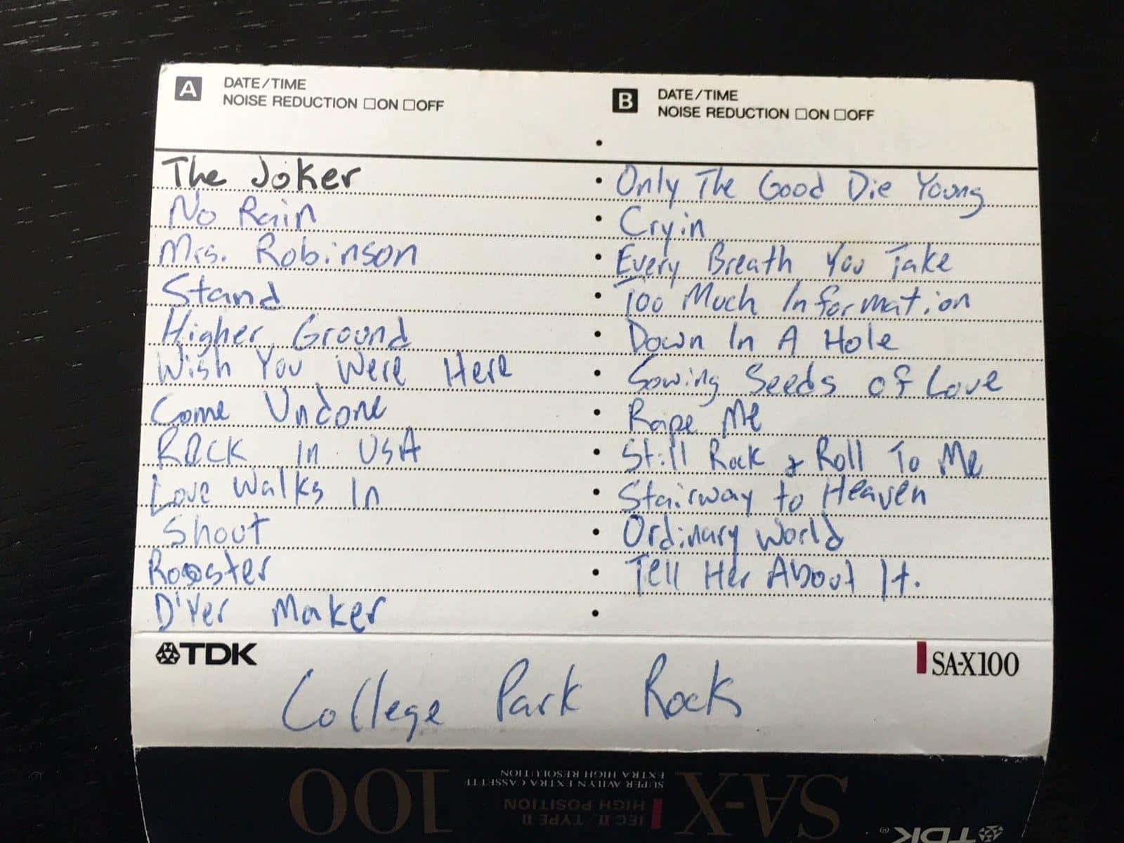 Mixtape 桌布 壁紙 — 在深色木質表面上，用藍色墨水在白色橫線卡式錄音帶標籤上手寫的歌曲列表。
