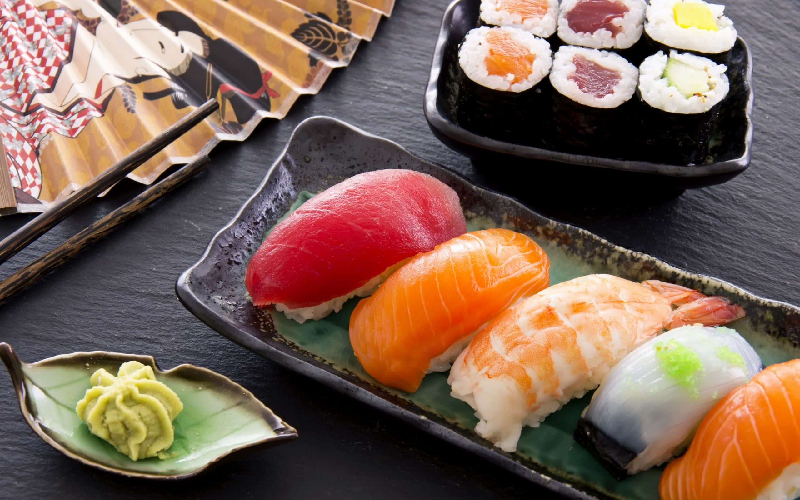 Sushi 桌布 壁紙 — 在深色石板表面上，新鮮壽司的鮮豔組合，包括鮪魚、鮭魚和蝦握壽司，以及卷壽司和芥末。