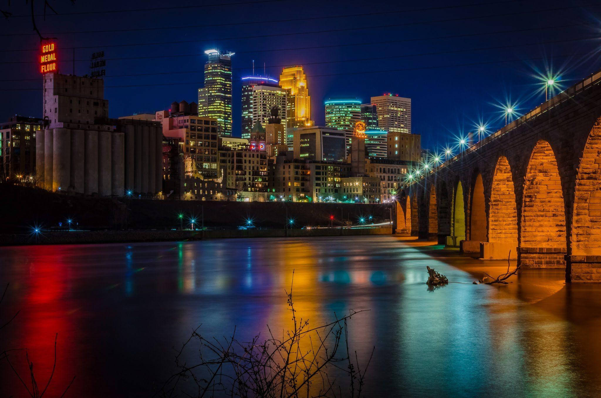Minnesota 桌布 壁紙 — 在深藍色天空下，一座石拱橋橫跨反射河面的活力夜景城市景觀。