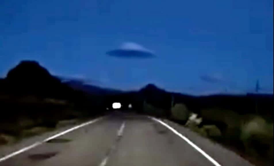 Road 桌布 壁紙 — 在深藍色天空下，一條模糊的道路在黃昏時分，發光的UFO懸浮在遠處山脈之上。