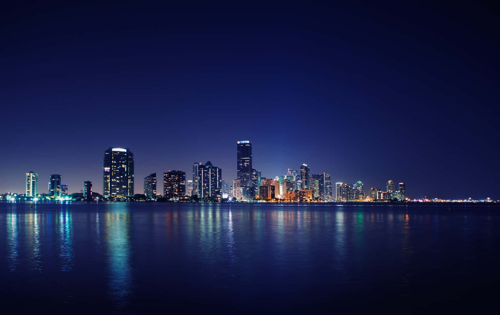 Miami 桌布 壁紙 — 在深藍色天空下，充滿活力的城市天際線在平靜的水面上倒映。