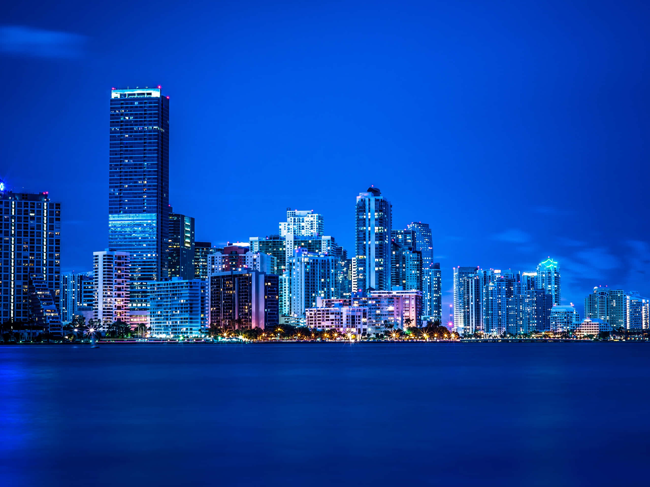 Miami 桌布 壁紙 — 在深藍色天空下，平靜水面上倒映著閃爍的摩天大樓，充滿活力的城市夜景。