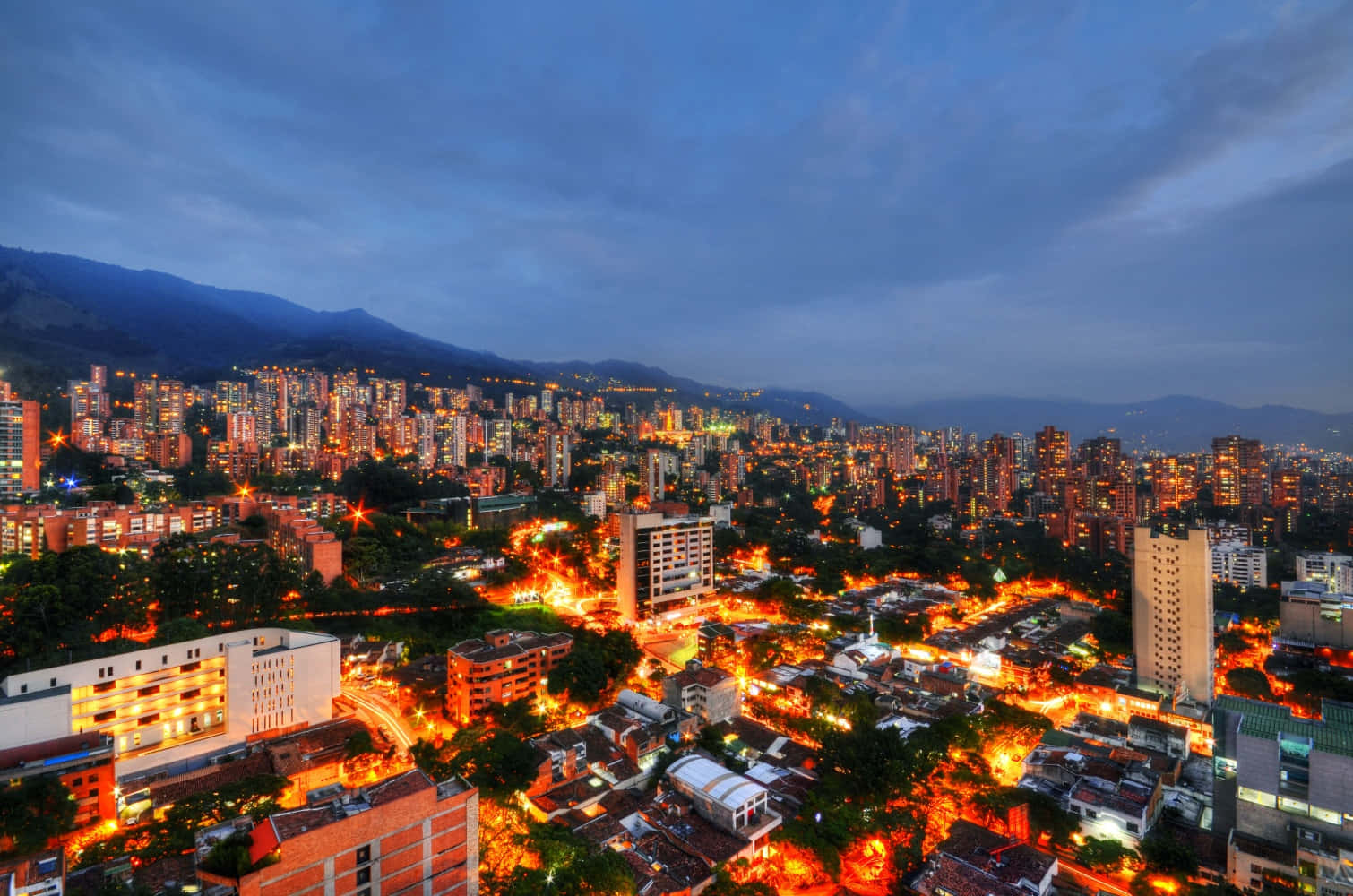 Colombia 桌布 壁紙 — 在深藍色天空下，廣闊的城市景觀在黃昏時分，溫暖的金色燈光從依偎在黑暗山脈旁的建築物中閃耀。