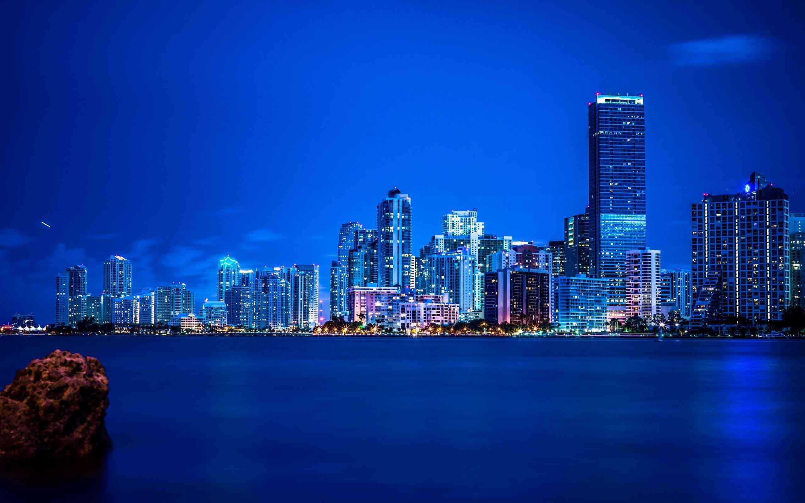 Miami 桌布 壁紙 — 在深藍色天空下，閃爍的摩天大樓倒映在平靜水面上，充滿活力的城市夜景。