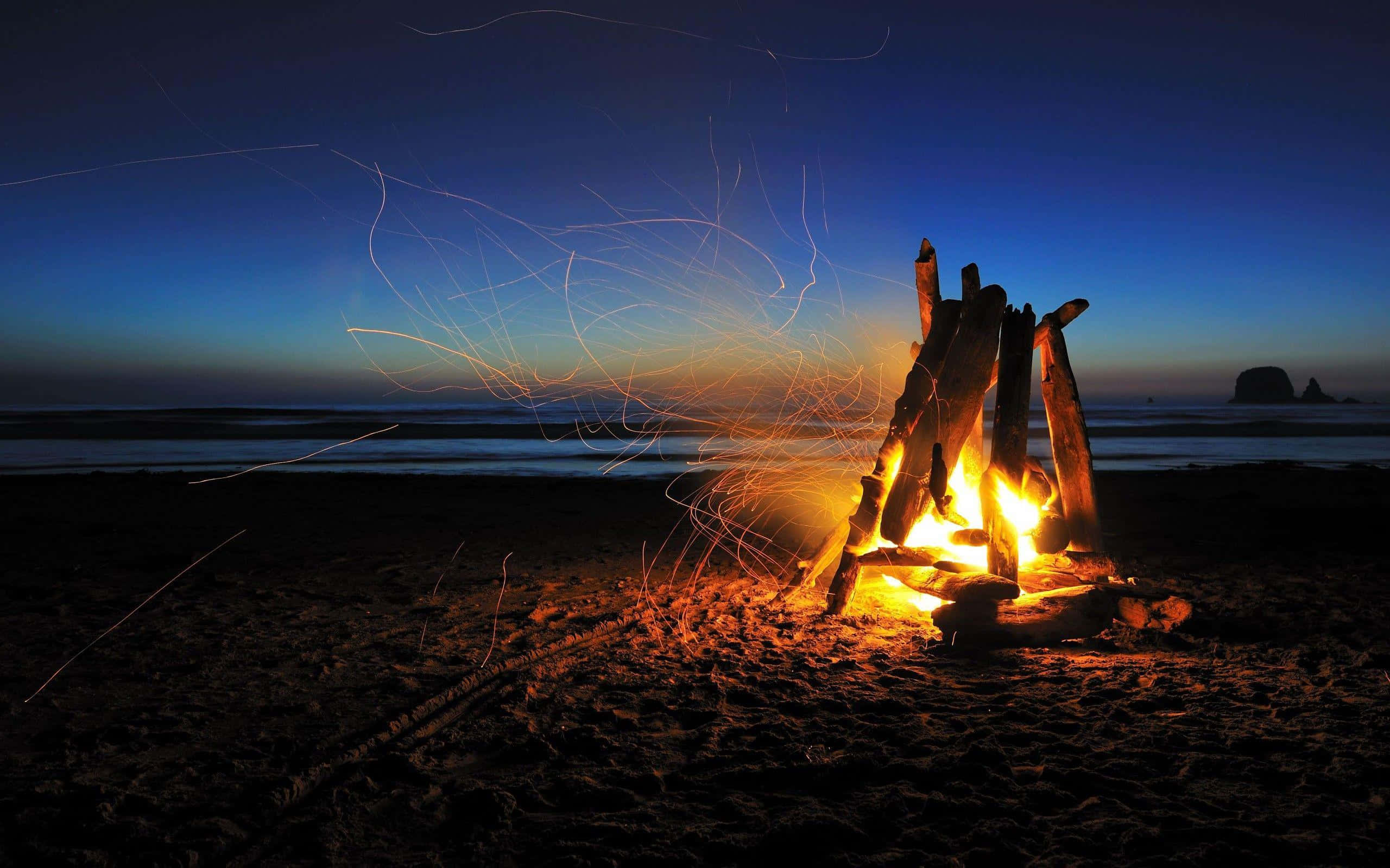 Camping 桌布 壁紙 — 在深藍色天空下，黑暗海灘上黃昏時分咆哮的營火，飛舞的火花和遠處的海石。