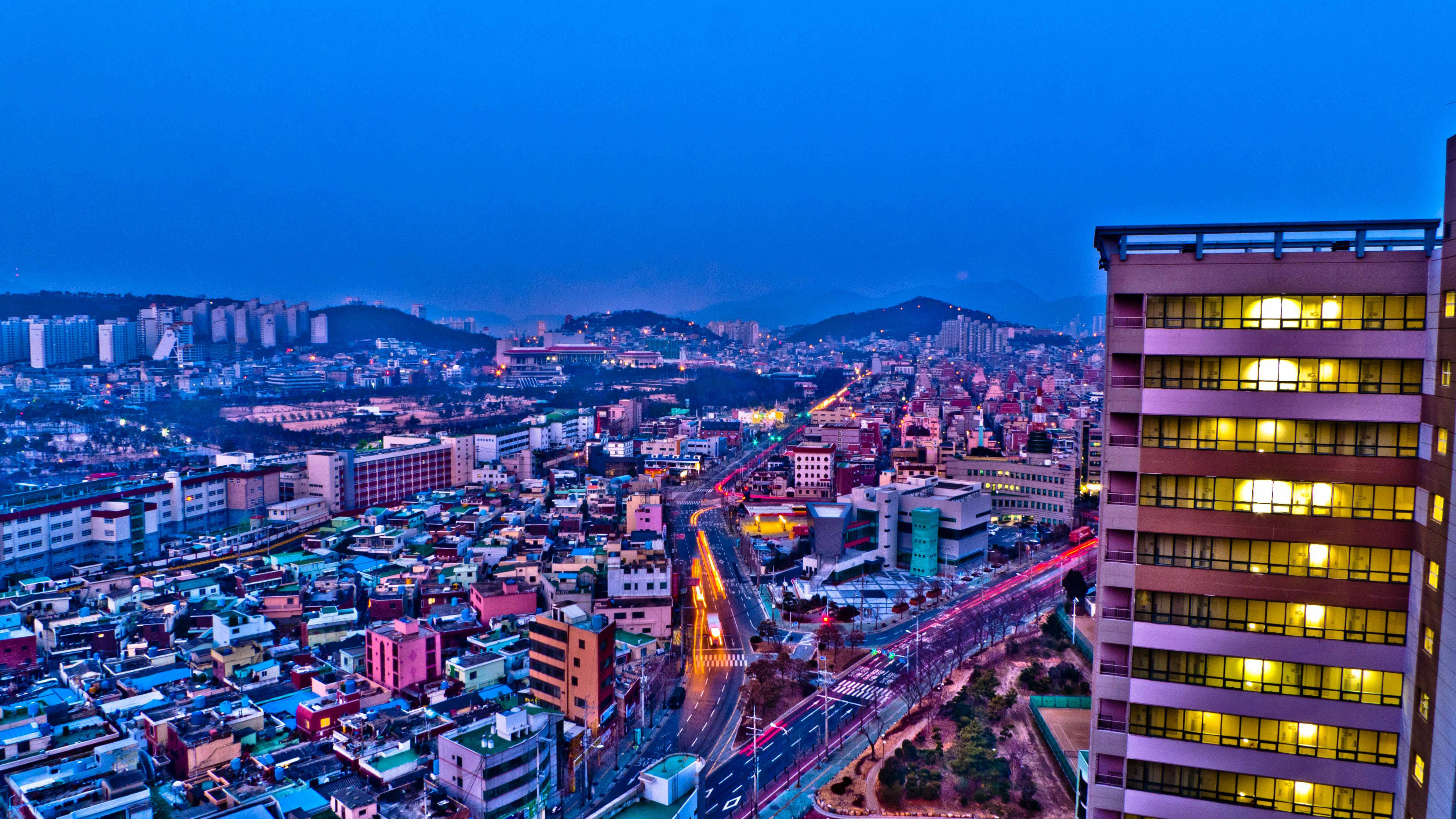 Busan 桌布 壁紙 — 在深藍色天空的襯托下，一個充滿活力的城市景觀，閃爍的窗戶和流動的交通燈光。