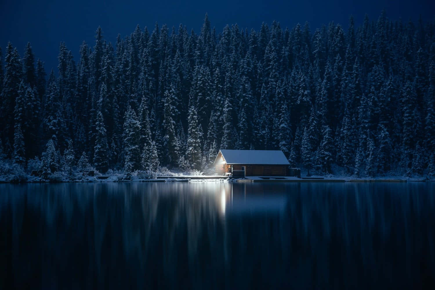 Cozy Winter Cabin 桌布 壁紙 — 在深藍色的冬夜天空下，一座孤獨的雪覆蓋小屋在結冰的湖邊溫暖地發光。