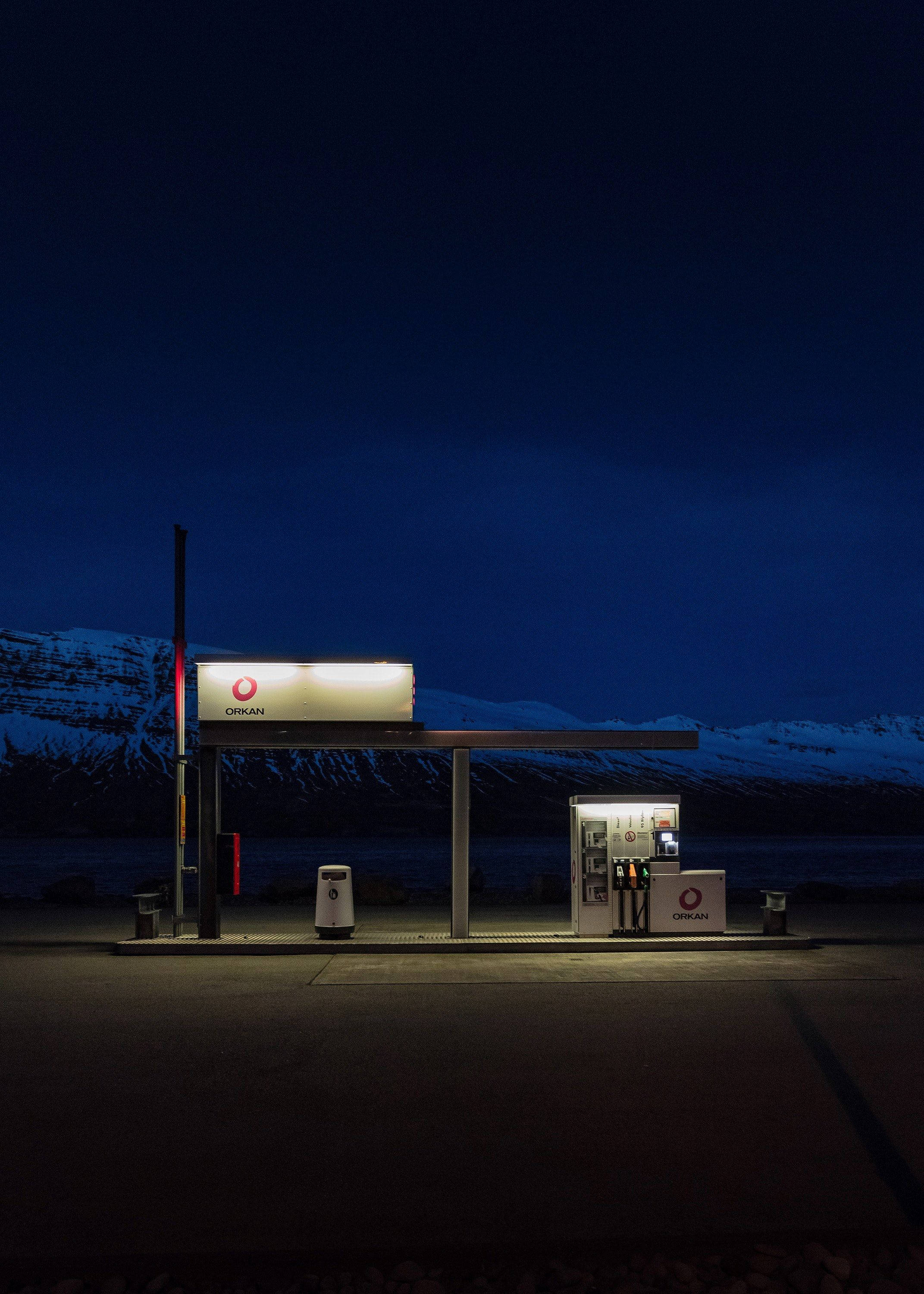 Gas Station 桌布 壁紙 — 在深藍色的天空下，一座孤獨的加油站夜晚亮著燈光，以積雪覆蓋的山脈為背景。