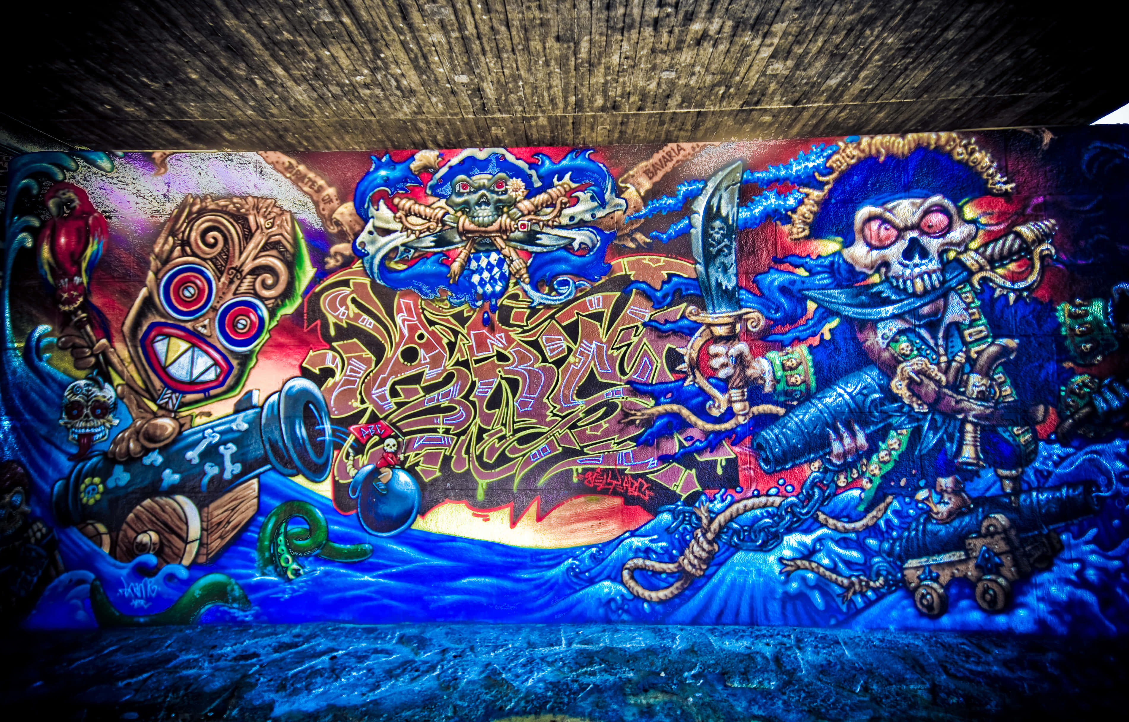 桌布 壁紙 — 在混凝土天橋下，描繪海盜頭骨和提基面具在暴風雨中海洋中戰鬥的鮮豔塗鴉壁畫。