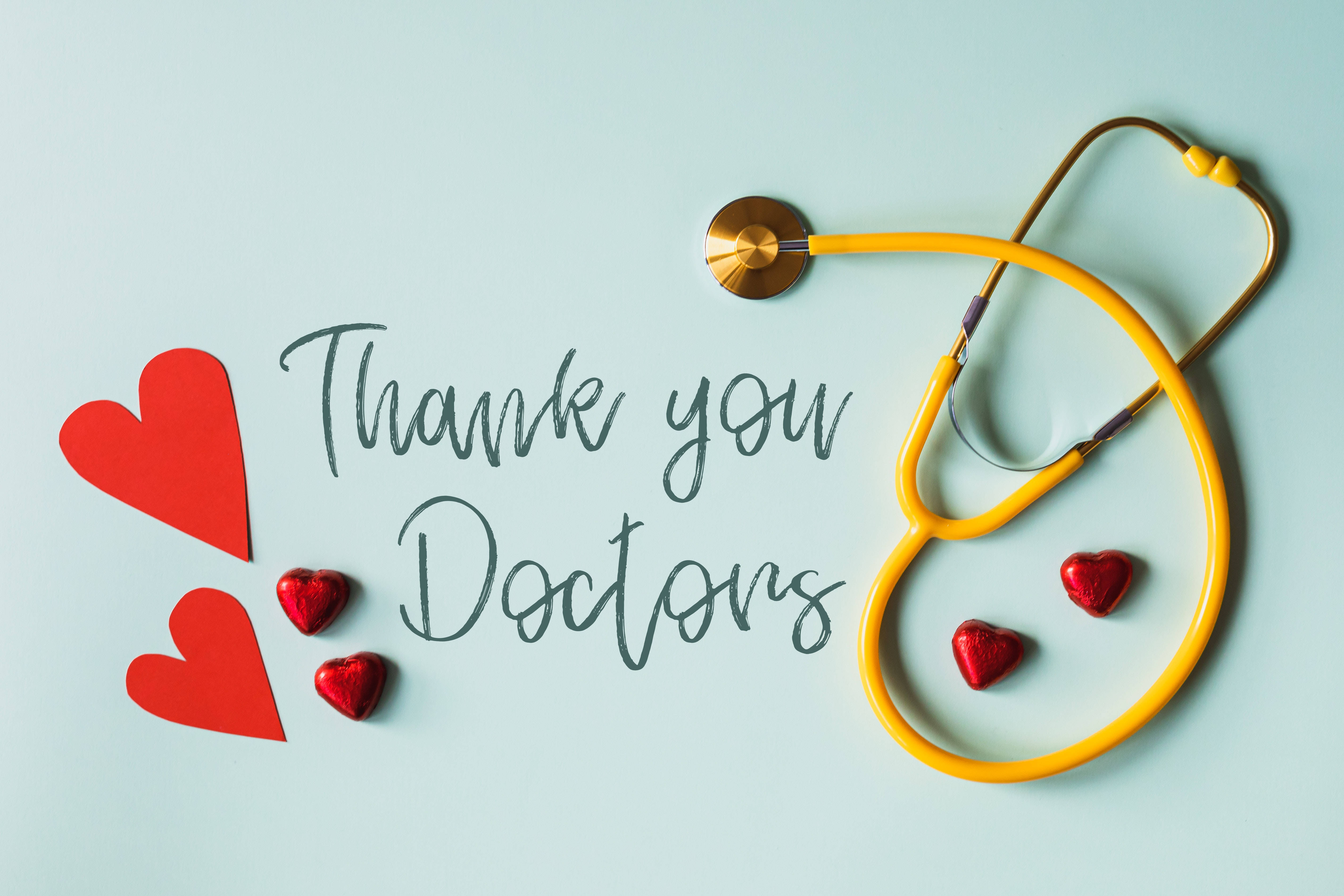 Doctors 桌布 壁紙 — 在淺藍色背景上，一個黃色聽診器和紅色心形圍繞著感謝醫生的手寫文字排列。
