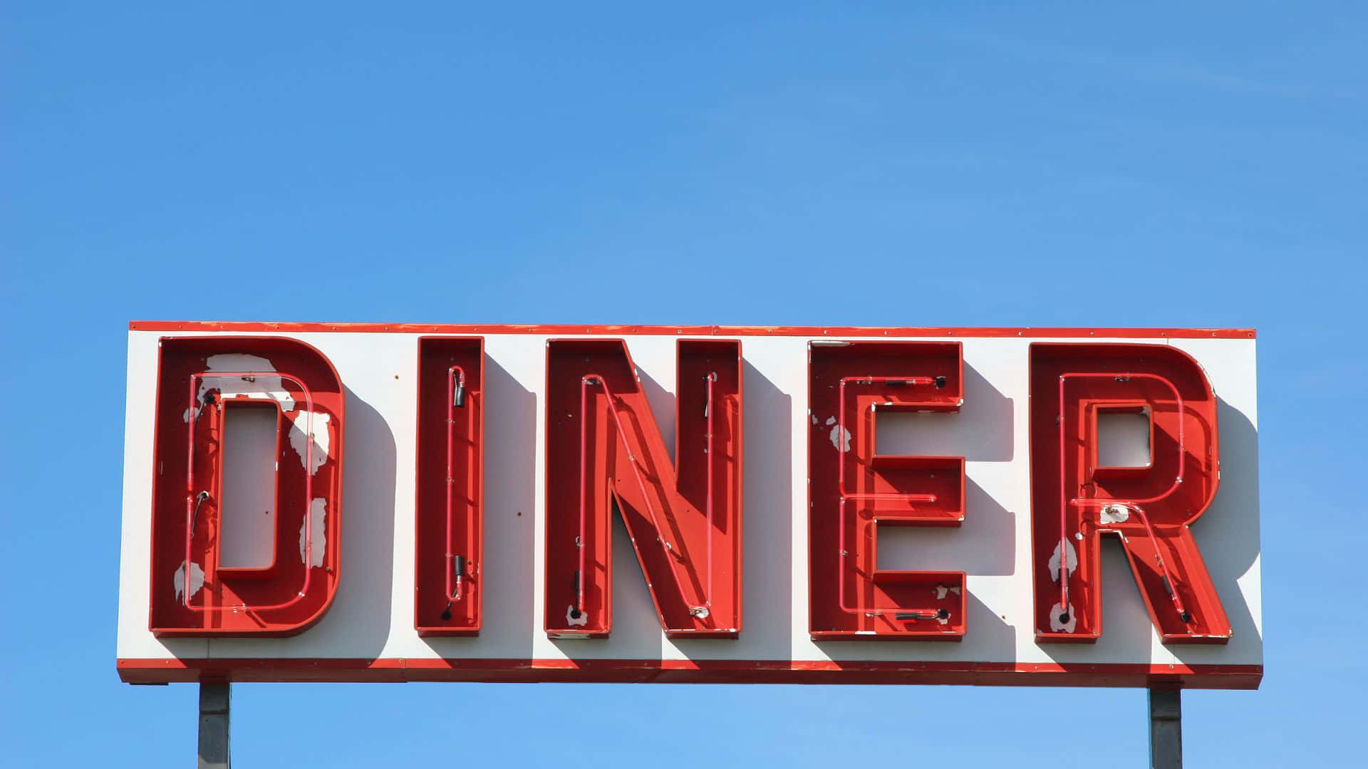 Diner 桌布 壁紙 — 在清澈的藍天下，一個風化的紅色霓虹燈招牌拼出 DINER。