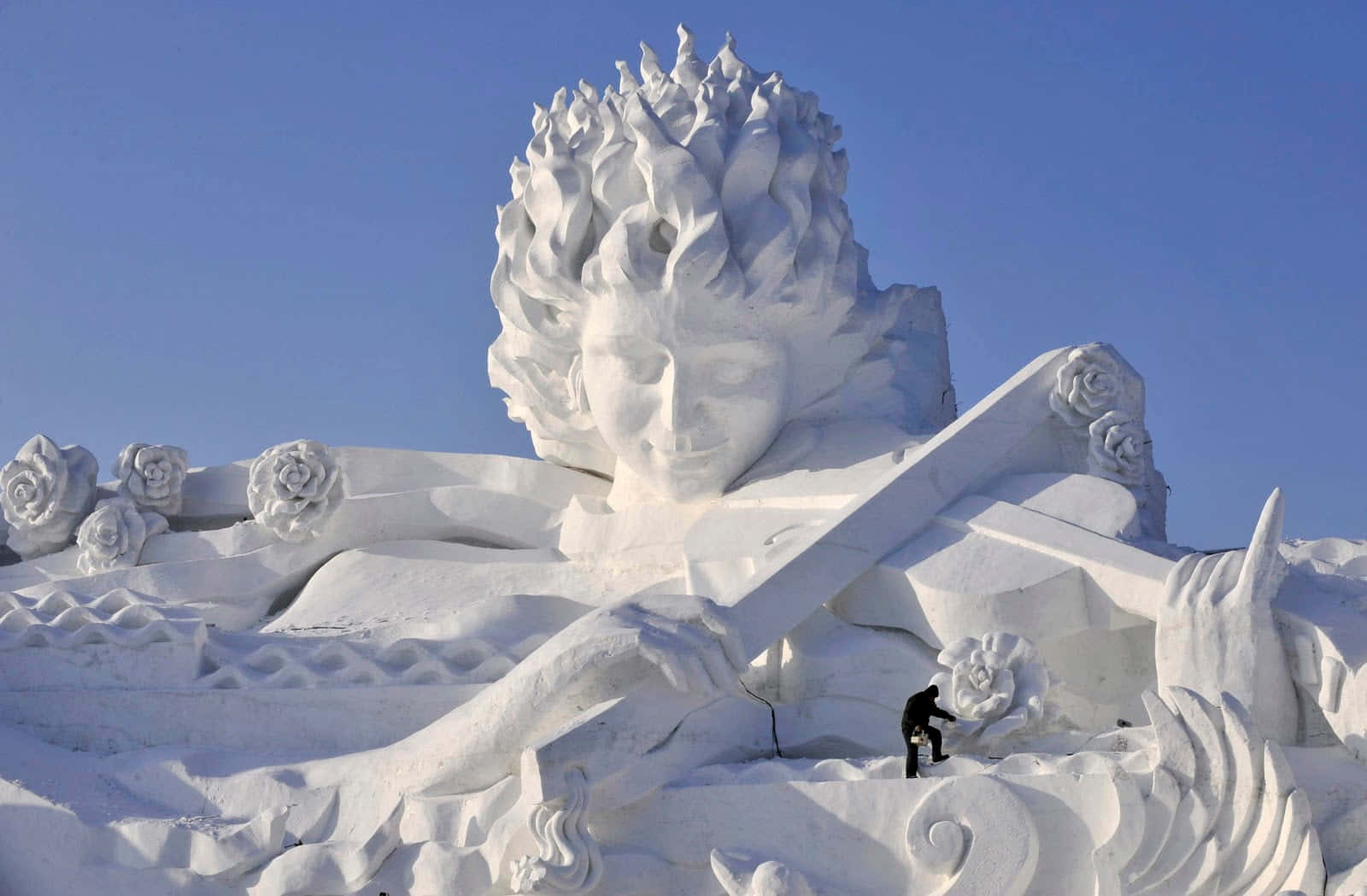 Ice Sculpture 桌布 壁紙 — 在清澈藍天下，一個帶有飄逸頭髮和玫瑰的寧靜面孔的巨大雪雕。