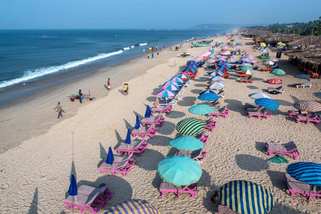 Goa Beach 桌布 壁紙 — 在清澈藍天下，粉紅色沙灘椅和彩色條紋遮陽傘排列的活力海灘鳥瞰圖。