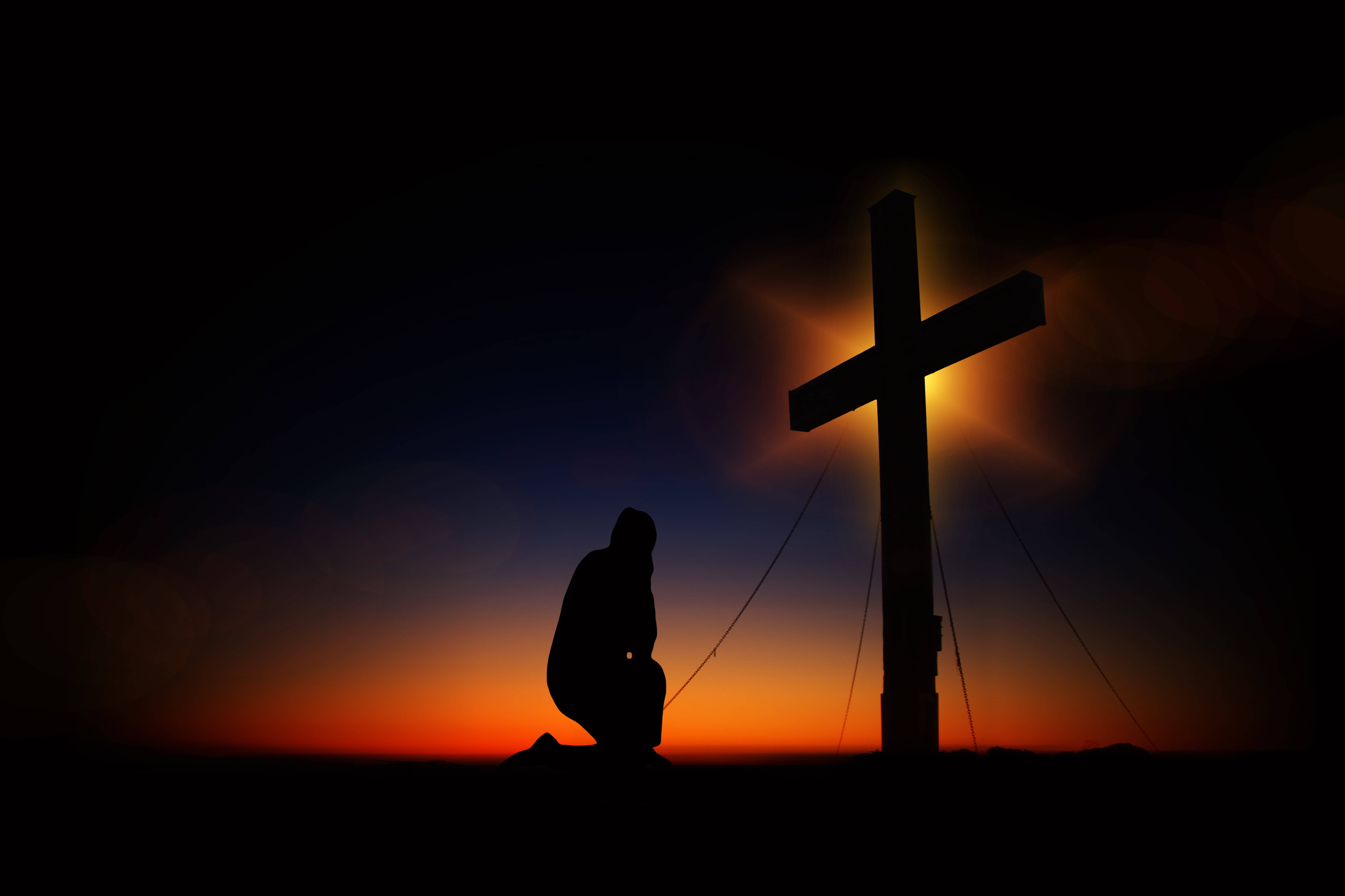 Christ 桌布 壁紙 — 在溫暖橙色和深藍色天空的黃昏時分，一個跪著祈禱的人物剪影，旁邊有一個發光的十字架。