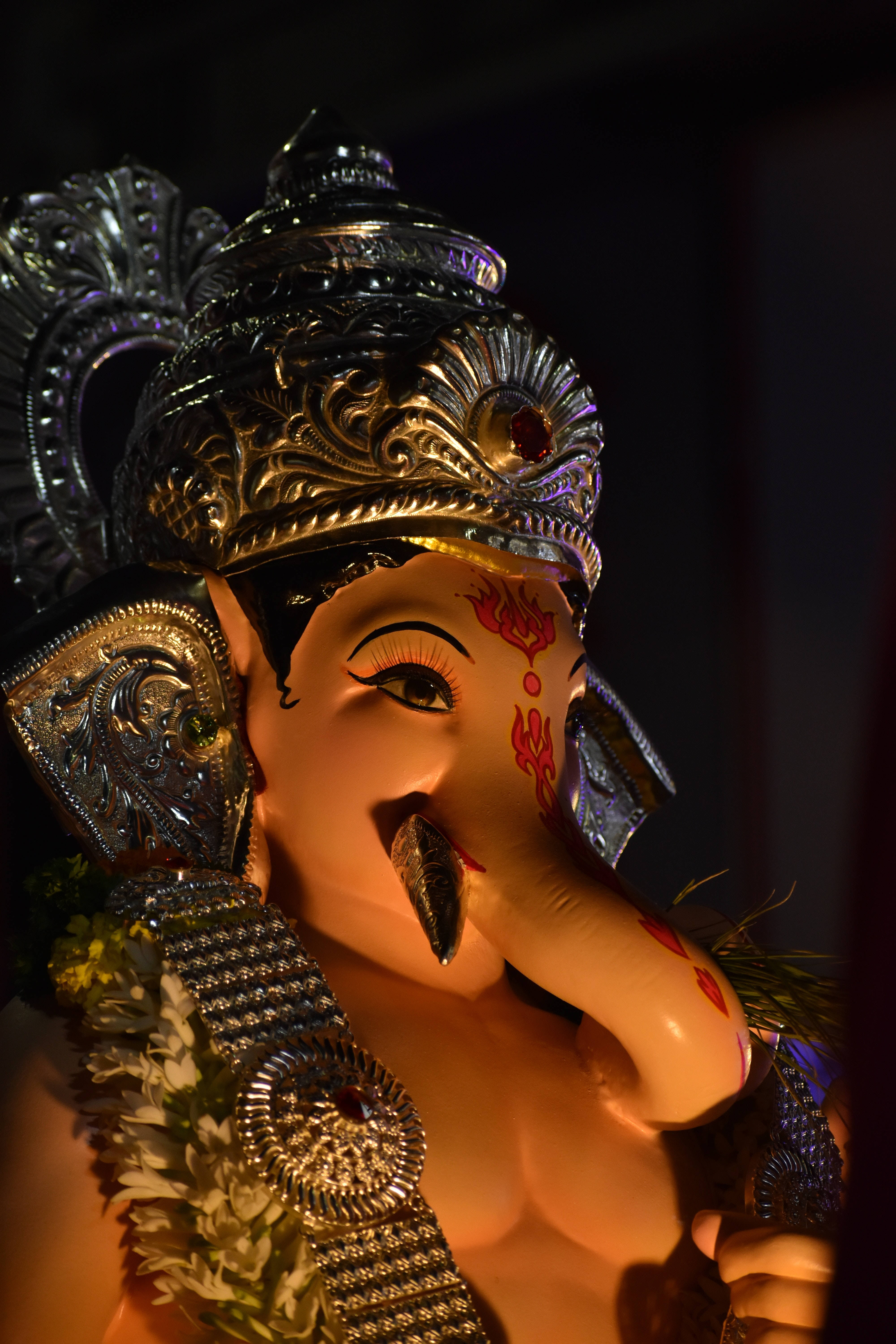 ganesh iPhone 桌布 壁紙 — 在溫暖燈光下，一尊裝飾華麗的象頭神雕像特寫，飾有精緻的銀飾和紅色花卉圖案。