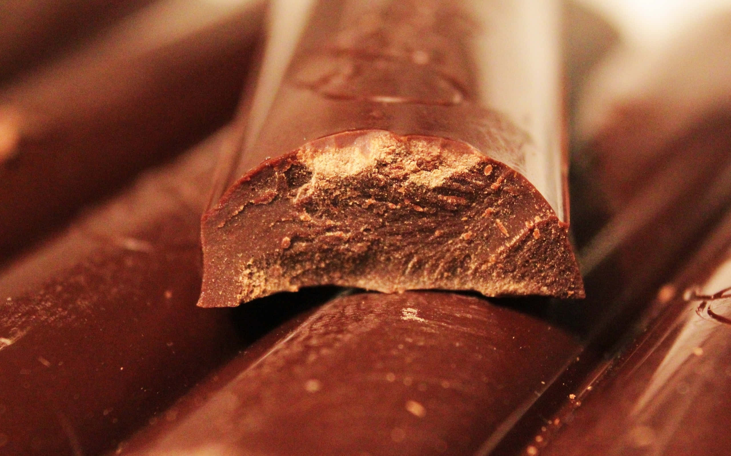 Brown Chocolate 桌布 壁紙 — 在溫暖燈光下，特寫一塊破碎的黑巧克力棒，展現其紋理豐富的內部。