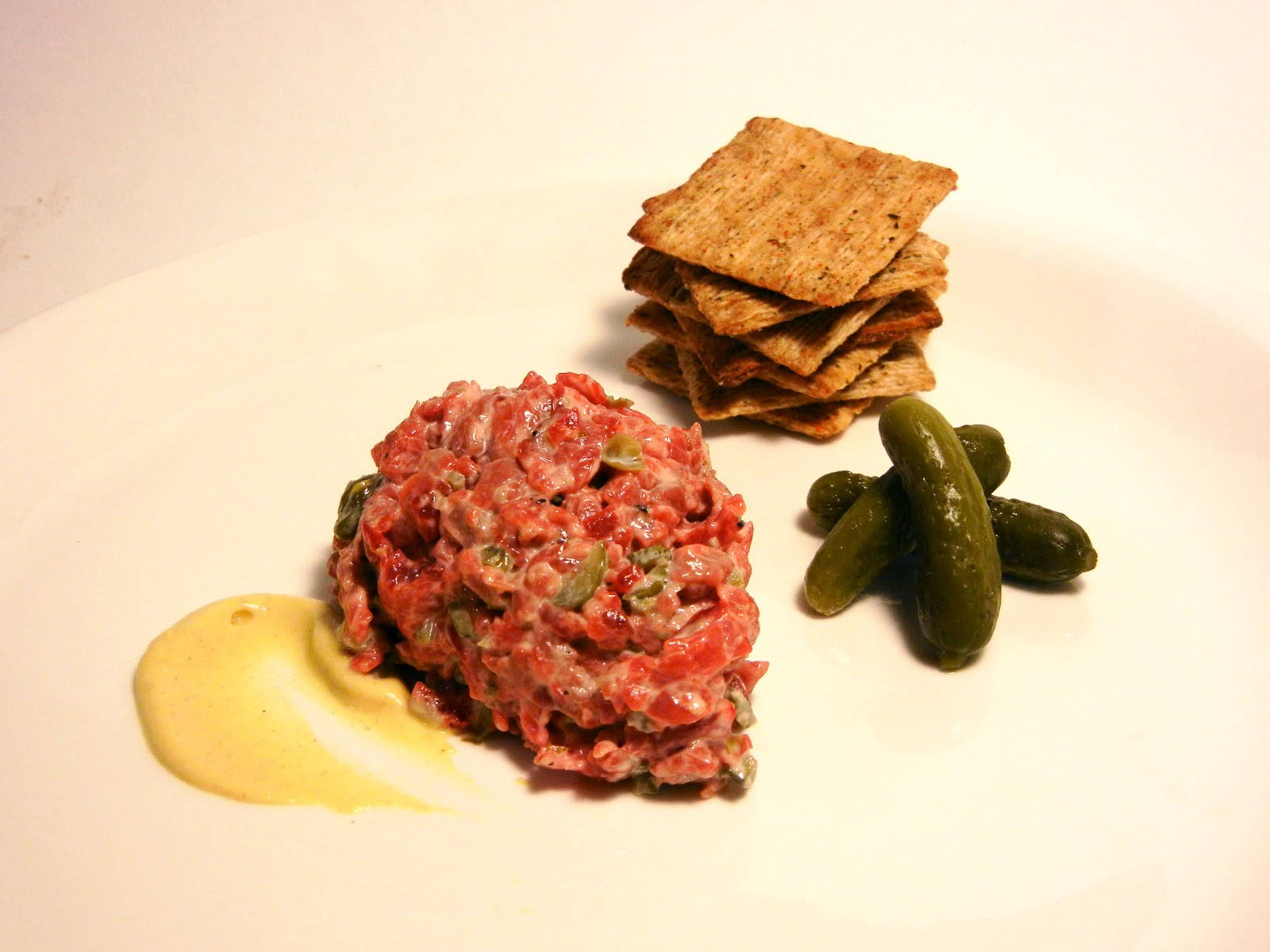 meat 桌布 壁紙 — 在溫暖燈光下，白色盤子上精心擺盤的牛肉塔塔山，搭配酸黃瓜和餅乾。