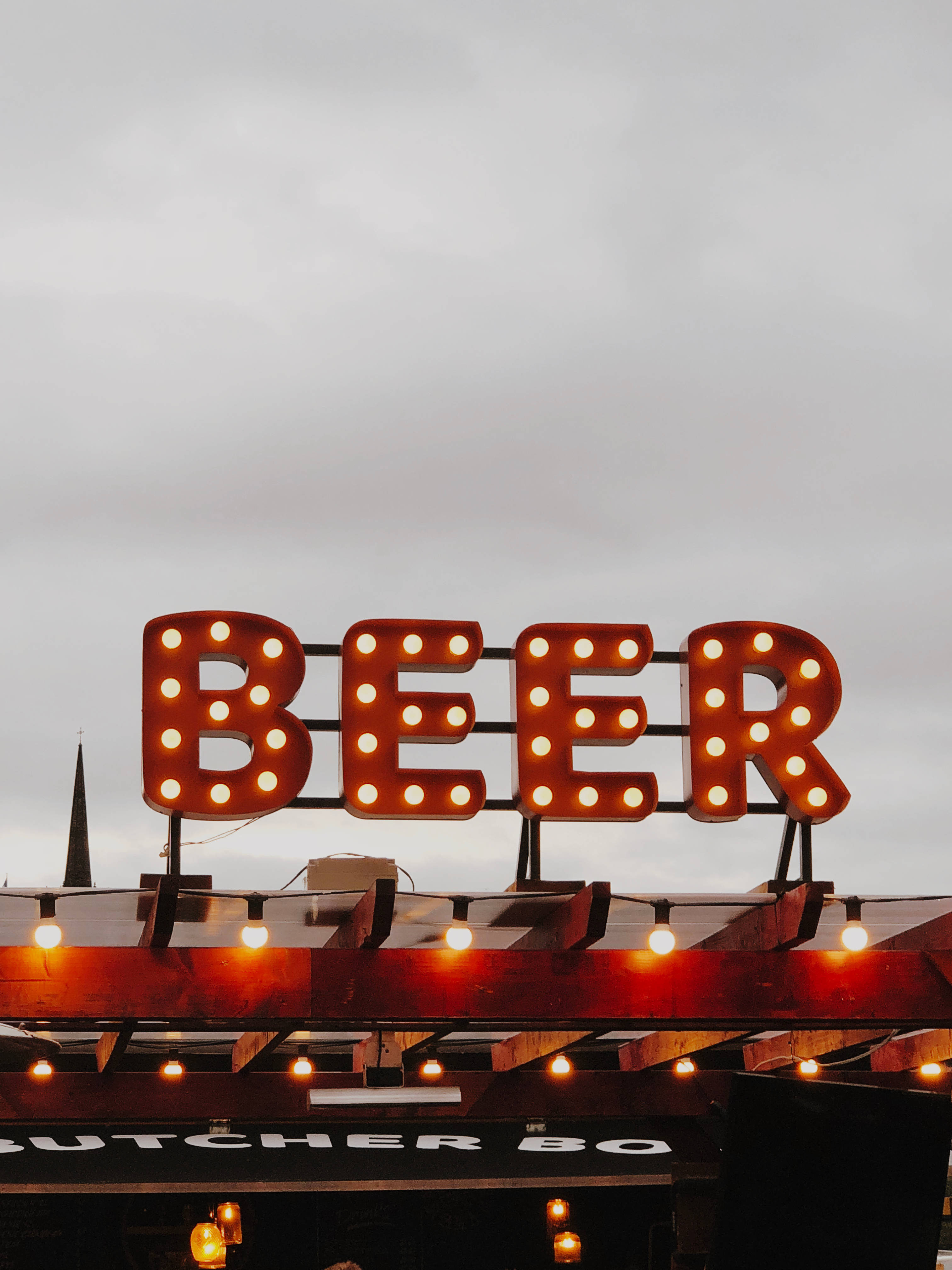 Beer 桌布 壁紙 — 在溫暖燈光下拼出BEER的發光霓虹招牌，背景為多雲天空，下方是質樸的木結構。
