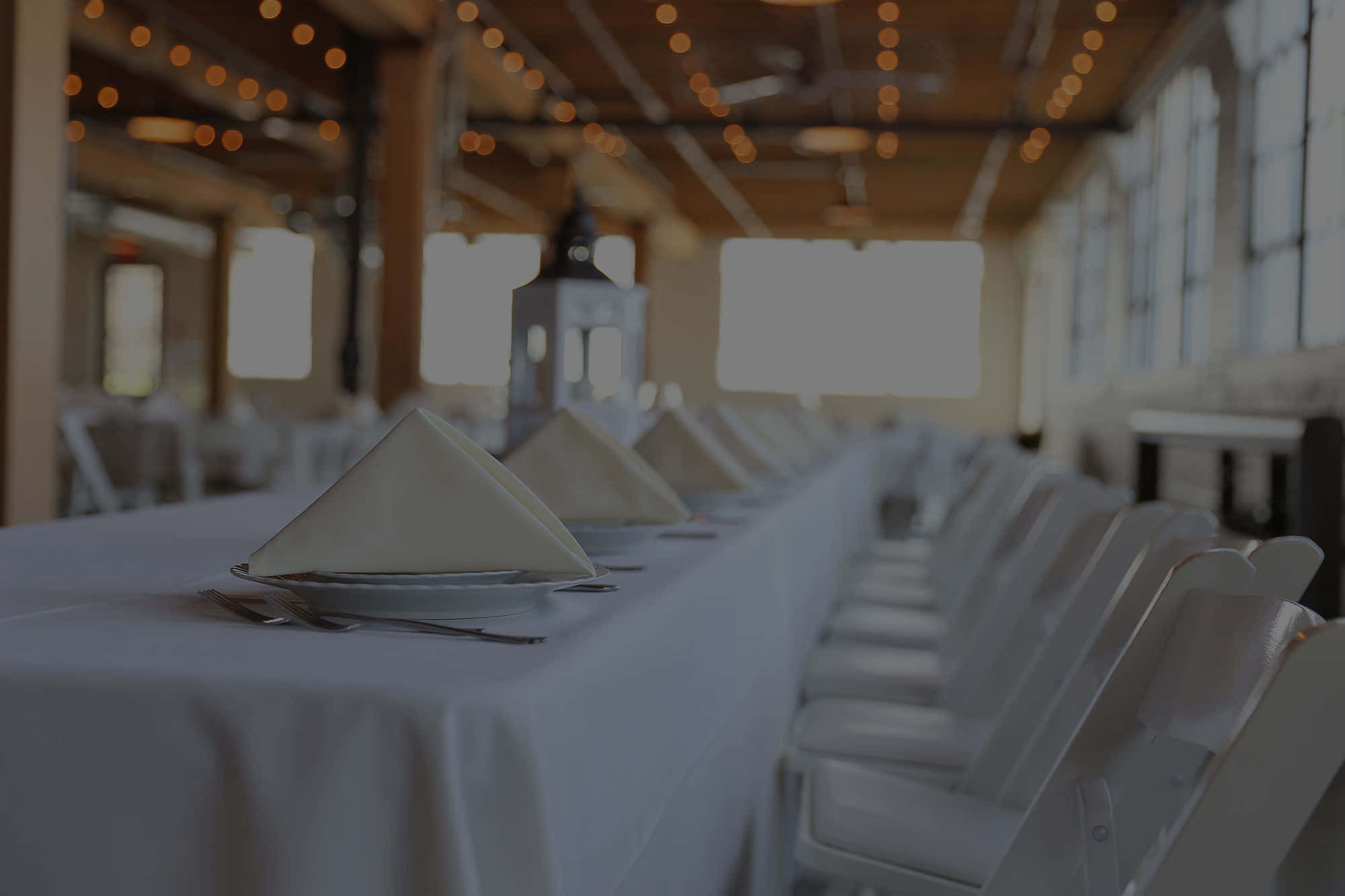 Hospitality 桌布 壁紙 — 在溫暖的串燈下，一個鄉村工業大廳裡擺放著鋪有白色亞麻布和折疊餐巾的長宴會桌。