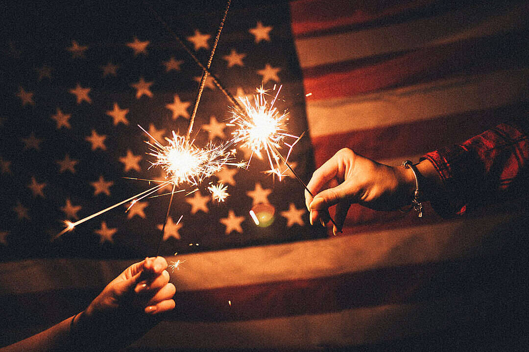 4th Of July 桌布 壁紙 — 在溫暖的夜間燈光下，兩隻手拿著閃閃發光的煙火，背景是柔和模糊的美國國旗。