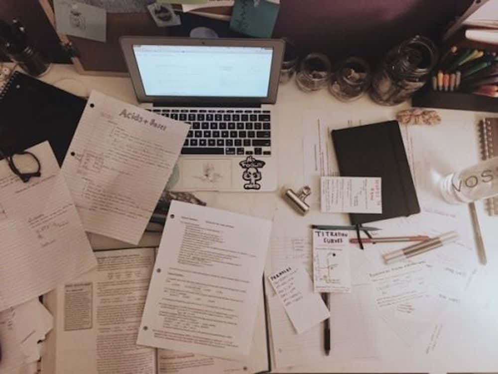書桌 桌布 壁紙 — 在溫暖的環境光線下，俯瞰一張雜亂的書桌，上面散落著化學筆記、筆記型電腦和文具。