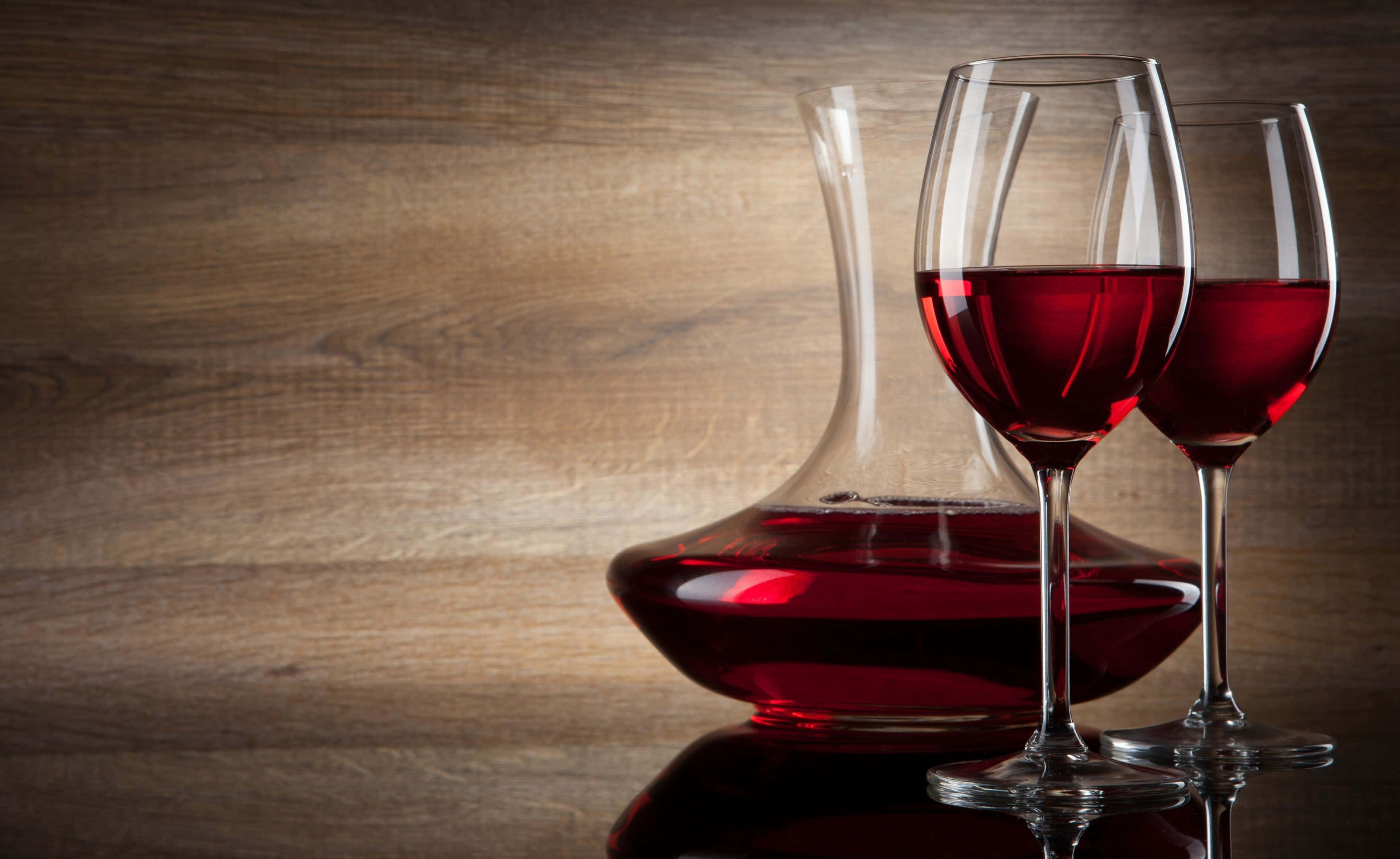 Red Wine Glass 桌布 壁紙 — 在溫暖的環境光線下，兩個玻璃杯和一個盛滿濃郁紅酒的醒酒器在拋光木質表面上閃閃發光。