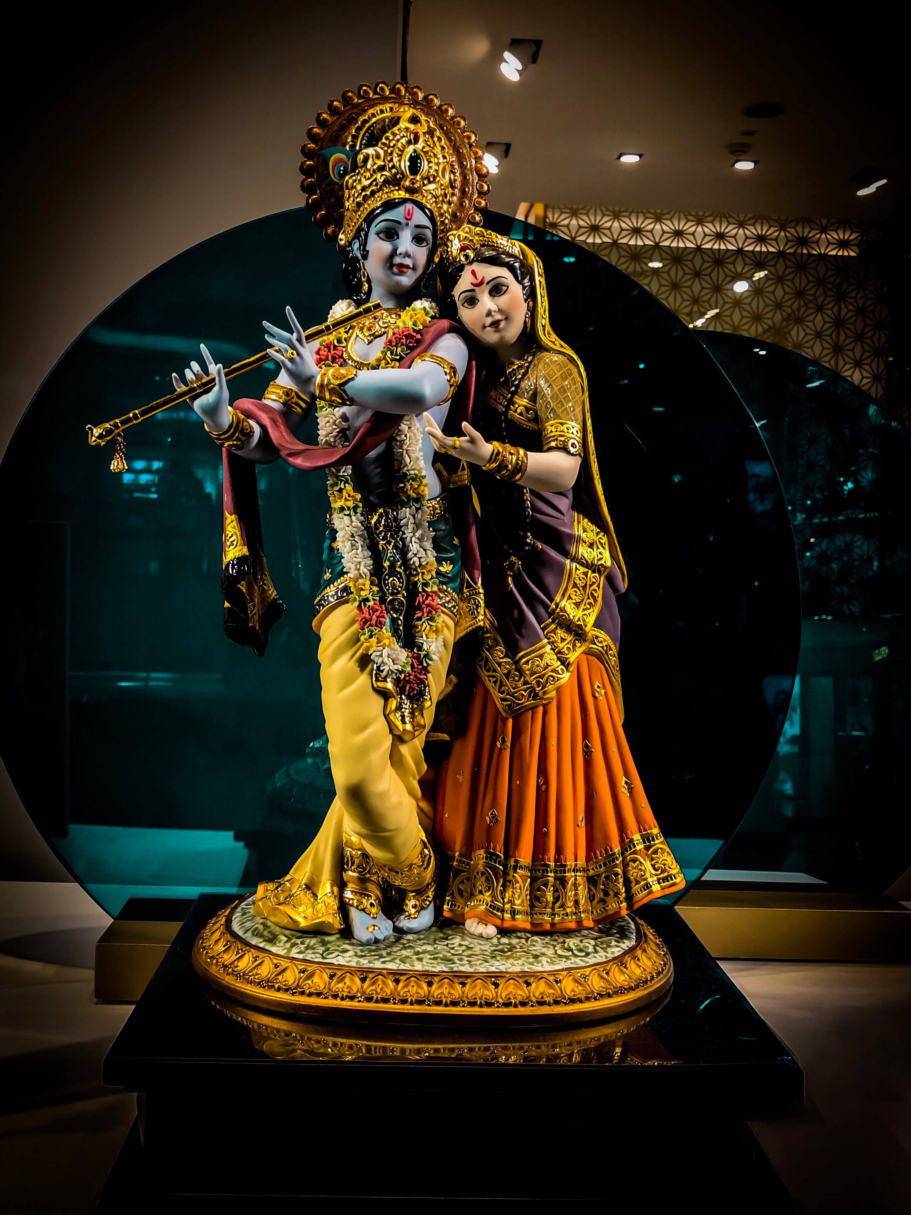 krishna iPhone 桌布 壁紙 — 在溫暖的畫廊燈光下，一尊金色的克里希納吹笛雕像，旁邊是穿著華麗傳統服飾的拉達。