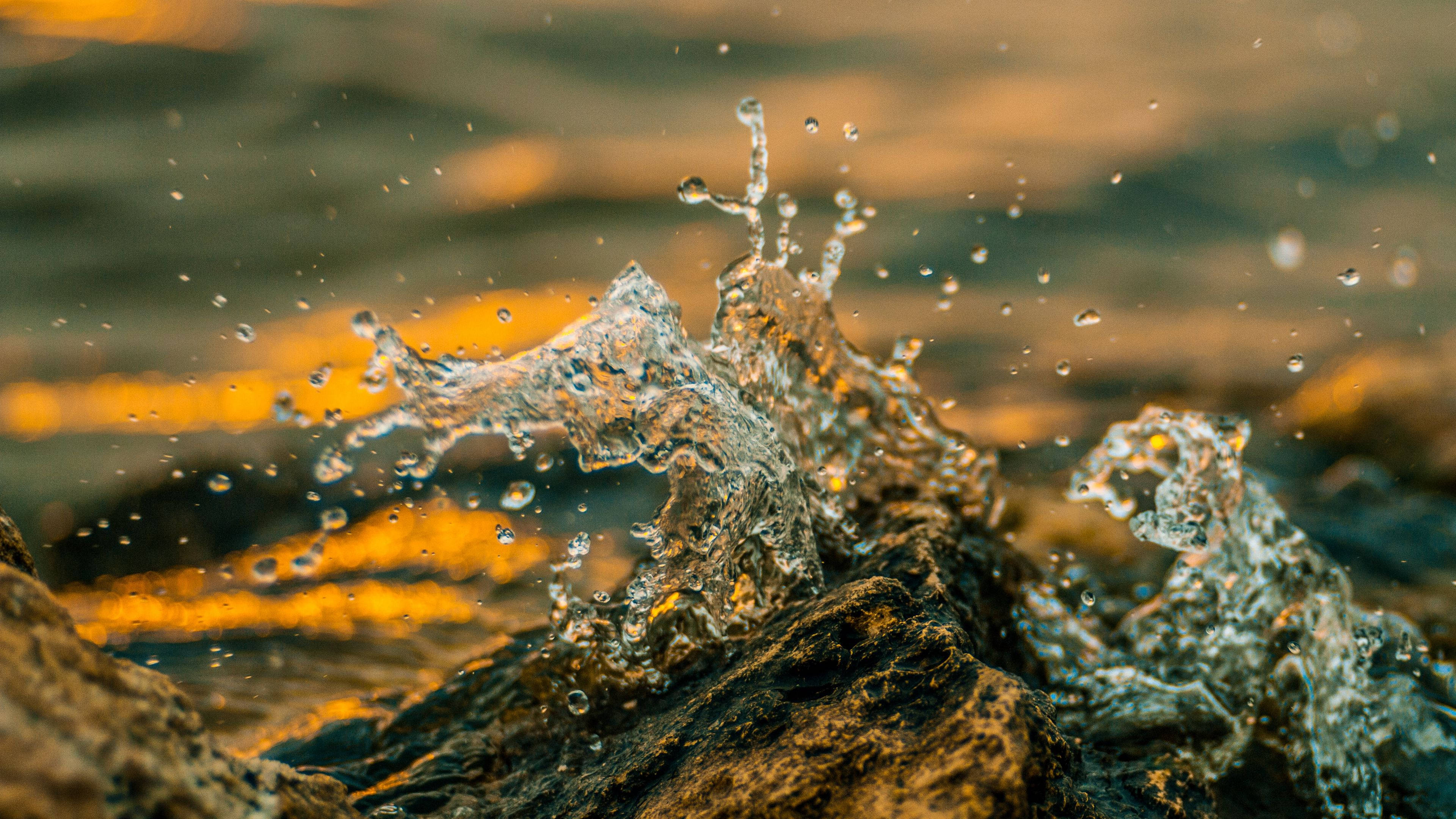 Water Splash 桌布 壁紙 — 在溫暖的金色日落背景下，水花從深色岩石中動態噴出。
