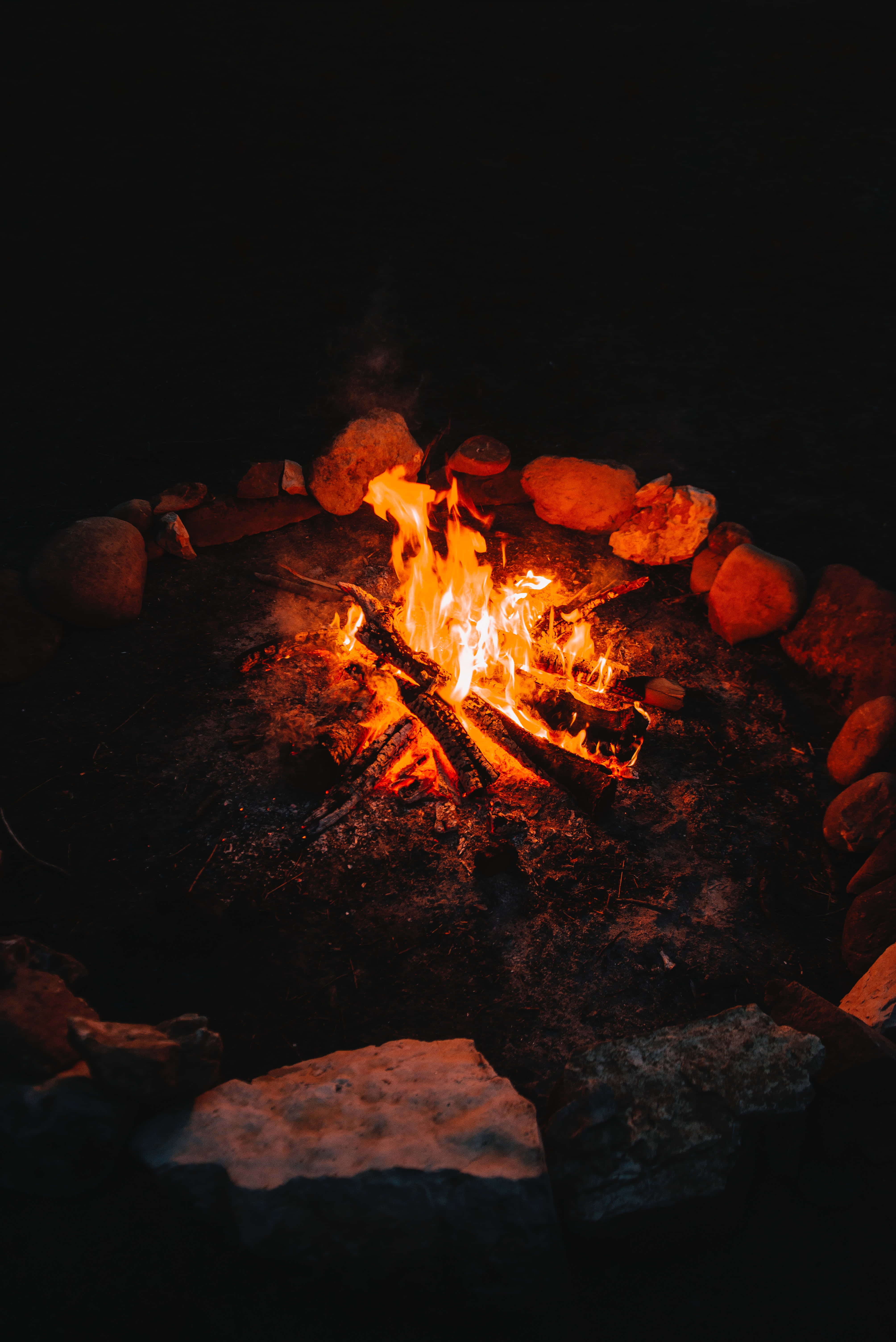 Bonfire 桌布 壁紙 — 在漆黑的夜空下，一個石圈內燃燒著旺盛的營火，將溫暖的橙色光芒投射在周圍的岩石上。