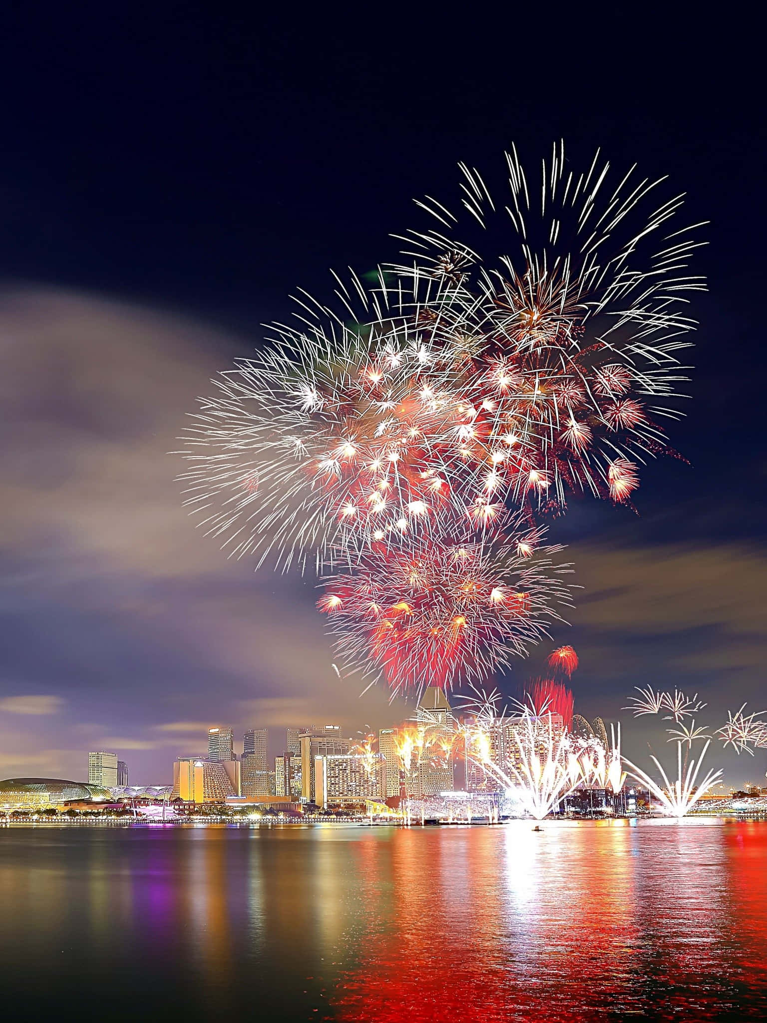 Fireworks 桌布 壁紙 — 在漆黑的夜空下，色彩繽紛的煙火在平靜水面倒映的城市天際線上綻放。