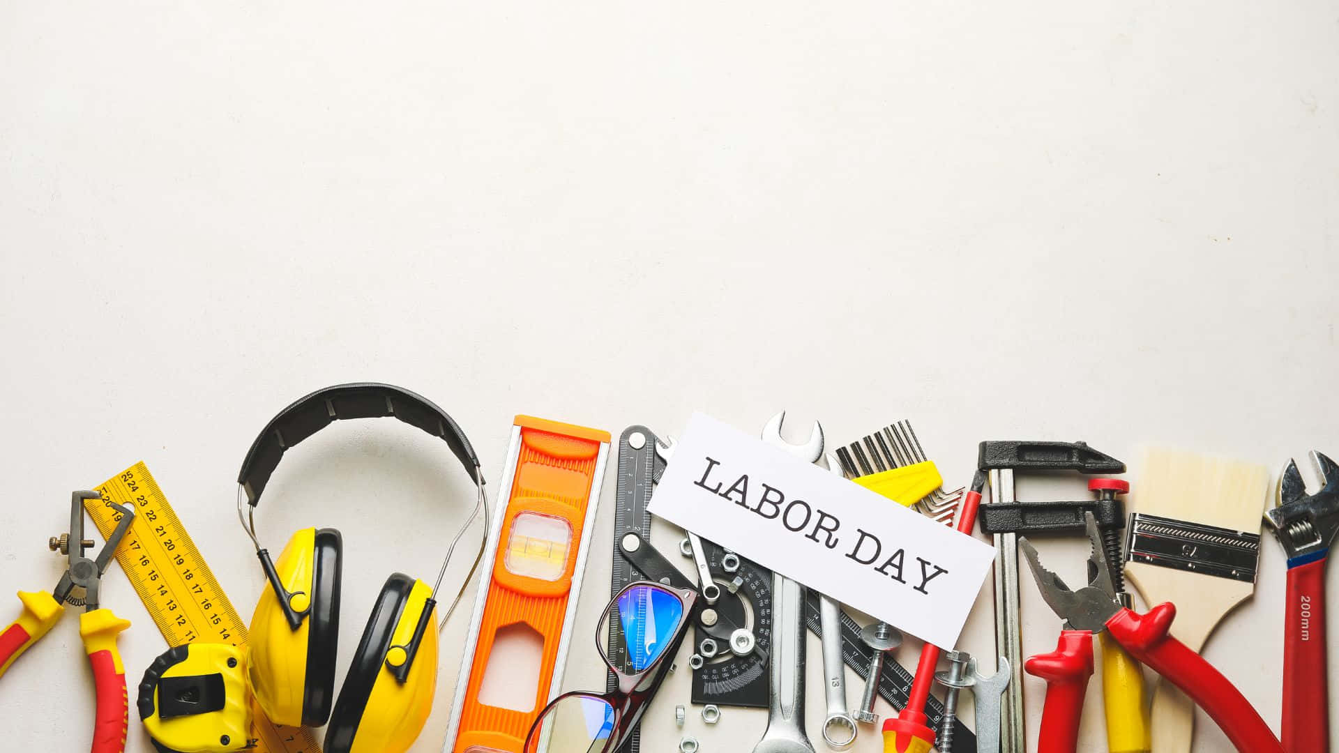 勞動節 桌布 壁紙 — 在潔白表面上，圍繞「LABOR DAY」標誌排列的彩色建築工具和安全裝備的平鋪佈局。