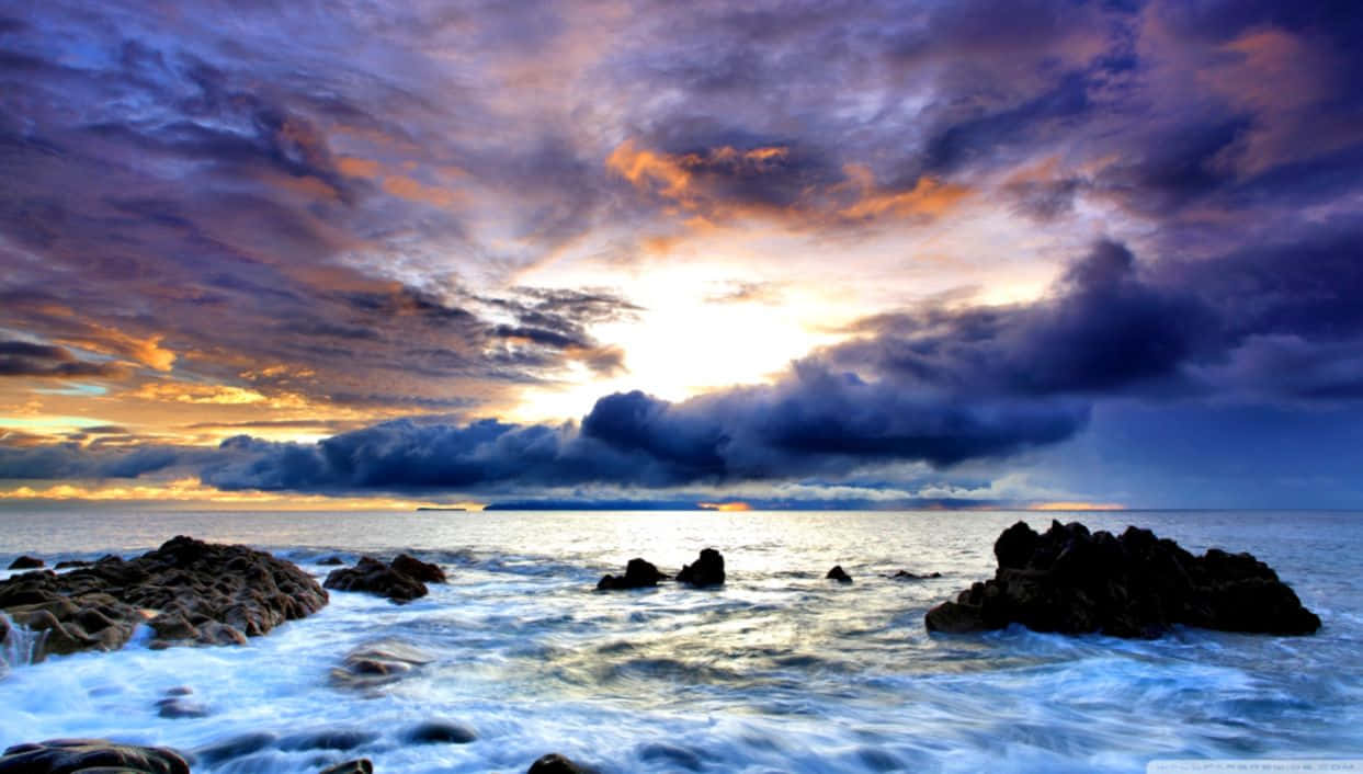 桌布 壁紙 — 在火橙色和深紫色雲層下，海洋上戲劇性的日落，海浪拍打著黑暗的岩石。