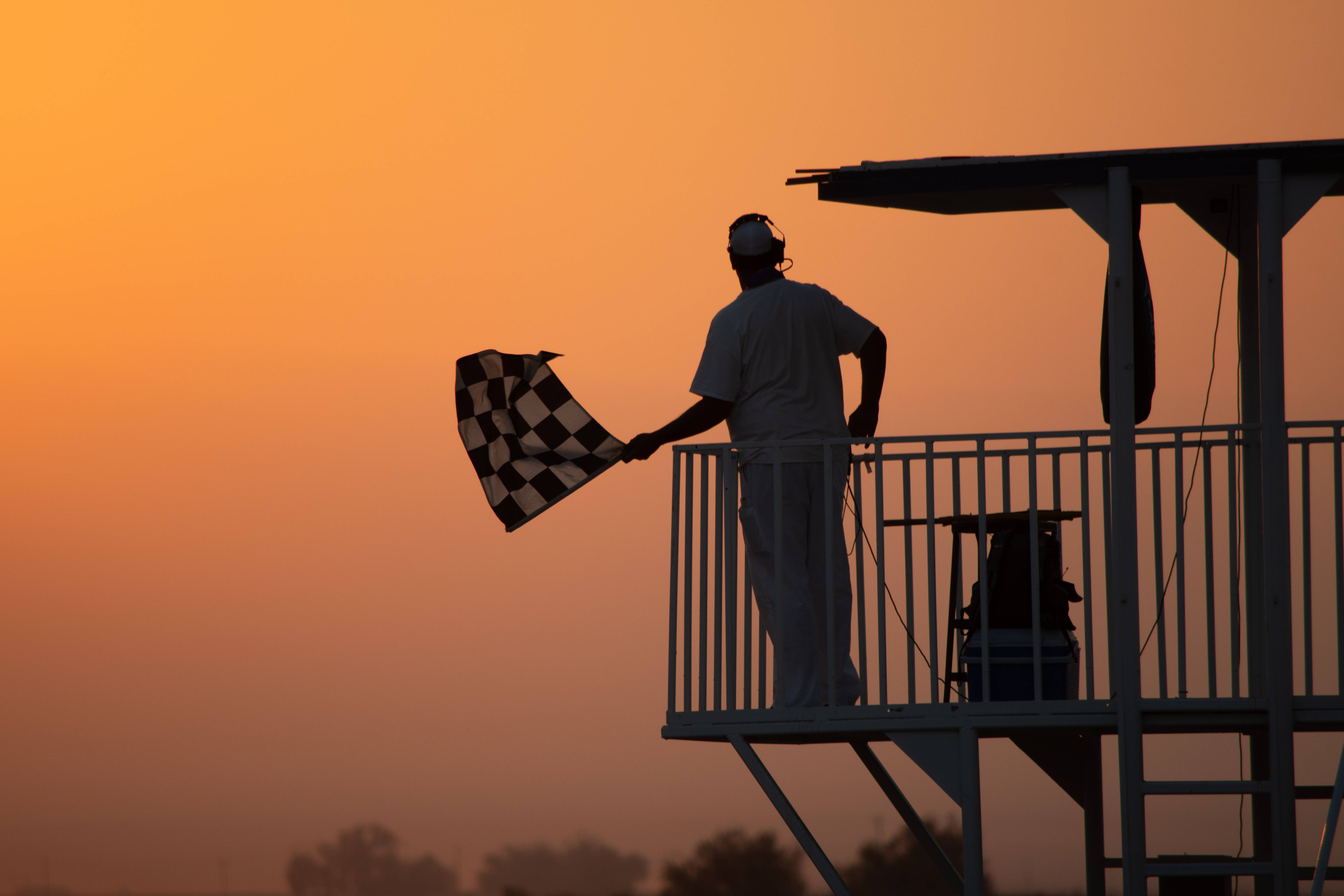 Checkered Flag 桌布 壁紙 — 在火熱橙色日落天空的背景下，一名賽事官員在塔頂揮舞格子旗的剪影。