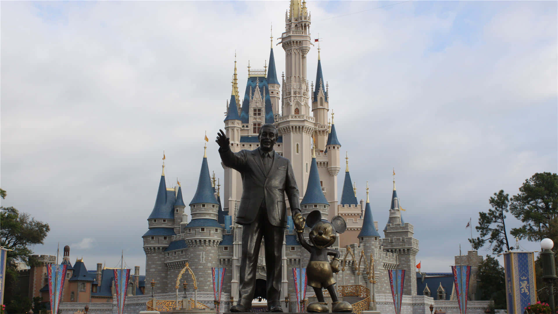Disney Castle 桌布 壁紙 — 在灰濛濛的天空下，灰姑娘城堡前，一座華特迪士尼的銅像正在揮手，旁邊是一個較小的米奇老鼠雕像。