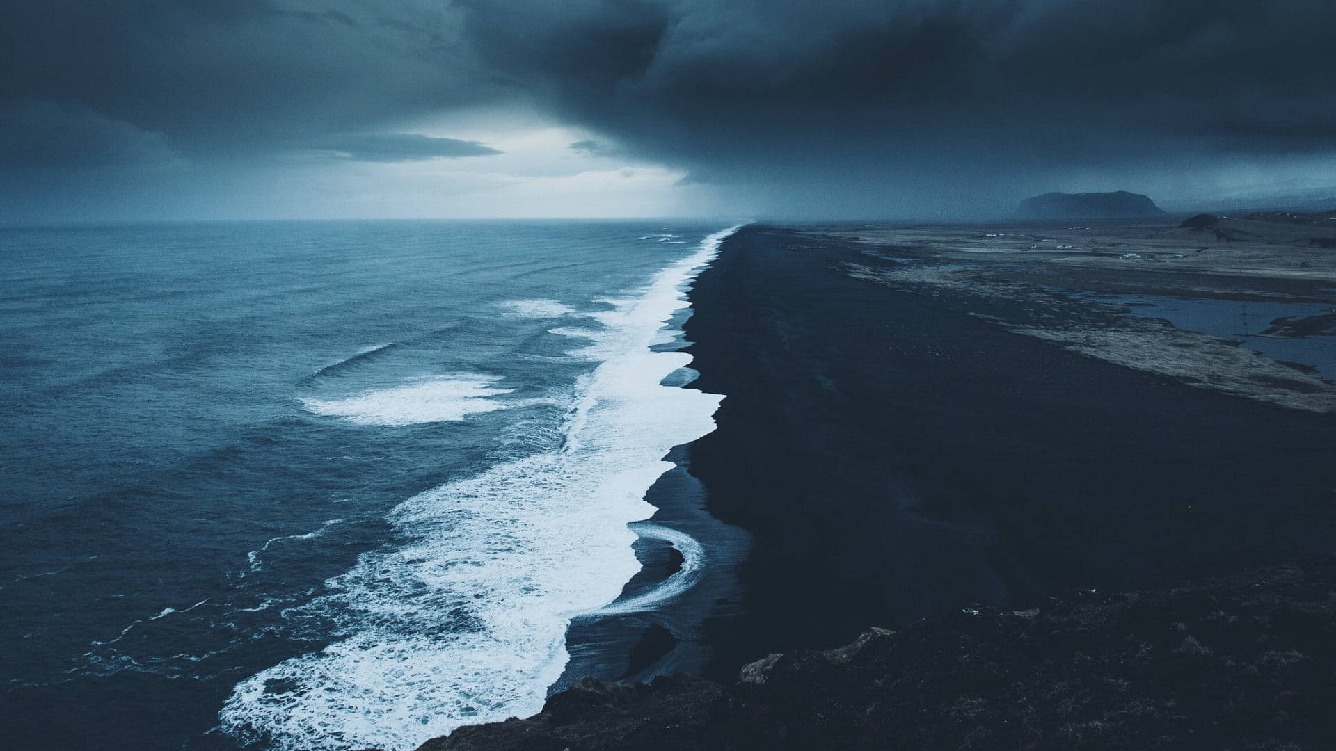 桌布 壁紙 — 在烏雲下，暴風雨海洋猛烈衝擊黑色火山海灘的戲劇性航拍視角。