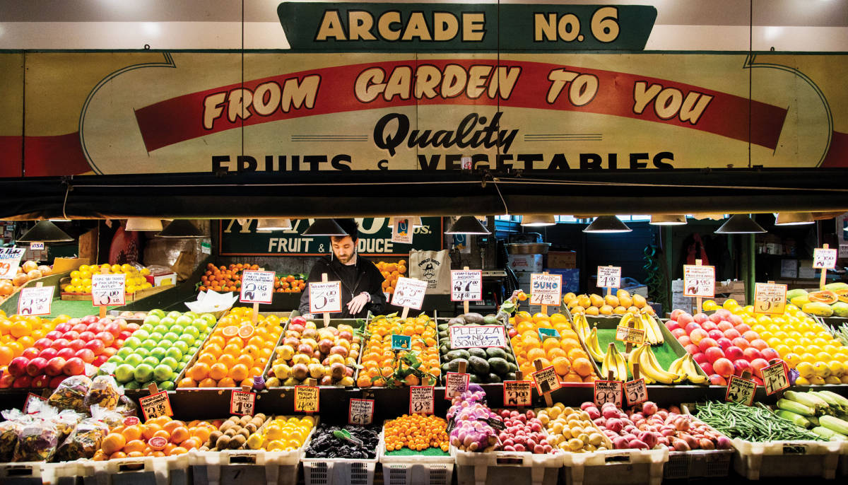 Pike Place Market 桌布 壁紙 — 在熙攘的市場中，復古拱廊招牌下色彩繽紛的水果和蔬菜堆滿了充滿活力的農產品攤位。