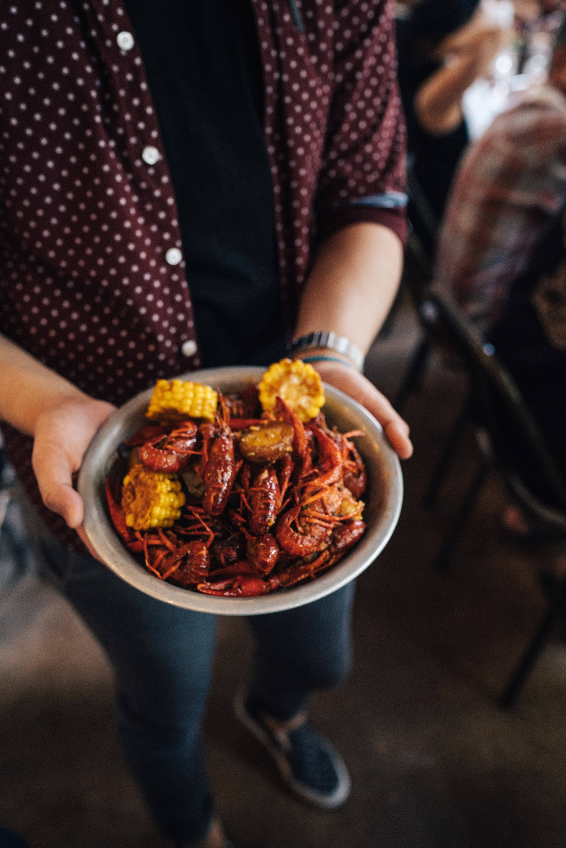 Crayfish 桌布 壁紙 — 在熱鬧的餐廳聚會中，有人拿著一碗冒著熱氣的小龍蝦和玉米棒。