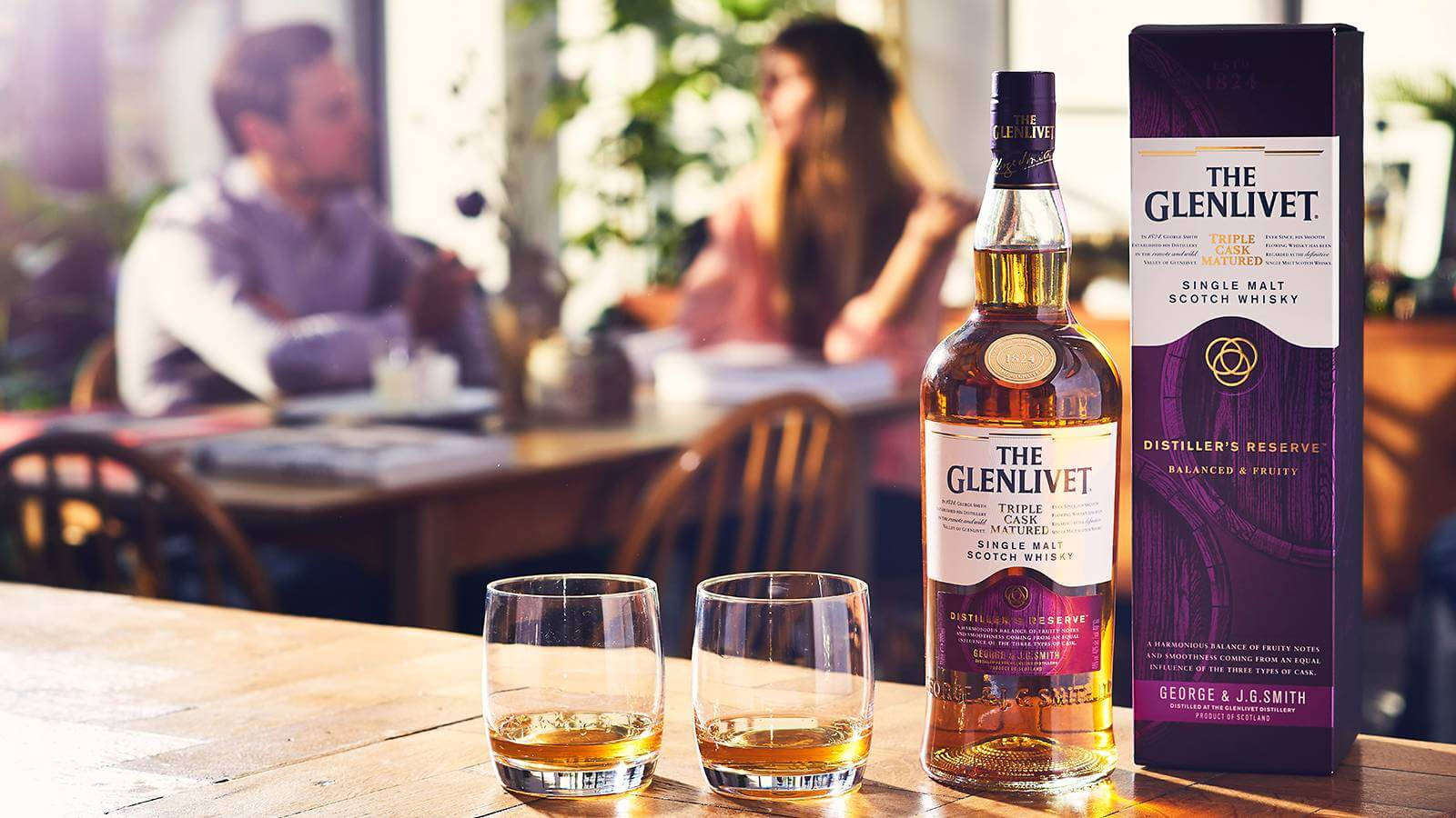 Box 桌布 壁紙 — 在燈光溫暖的餐廳裡，一瓶The Glenlivet Distiller's Reserve威士忌和兩個酒杯放在木桌上，背景有模糊的顧客。