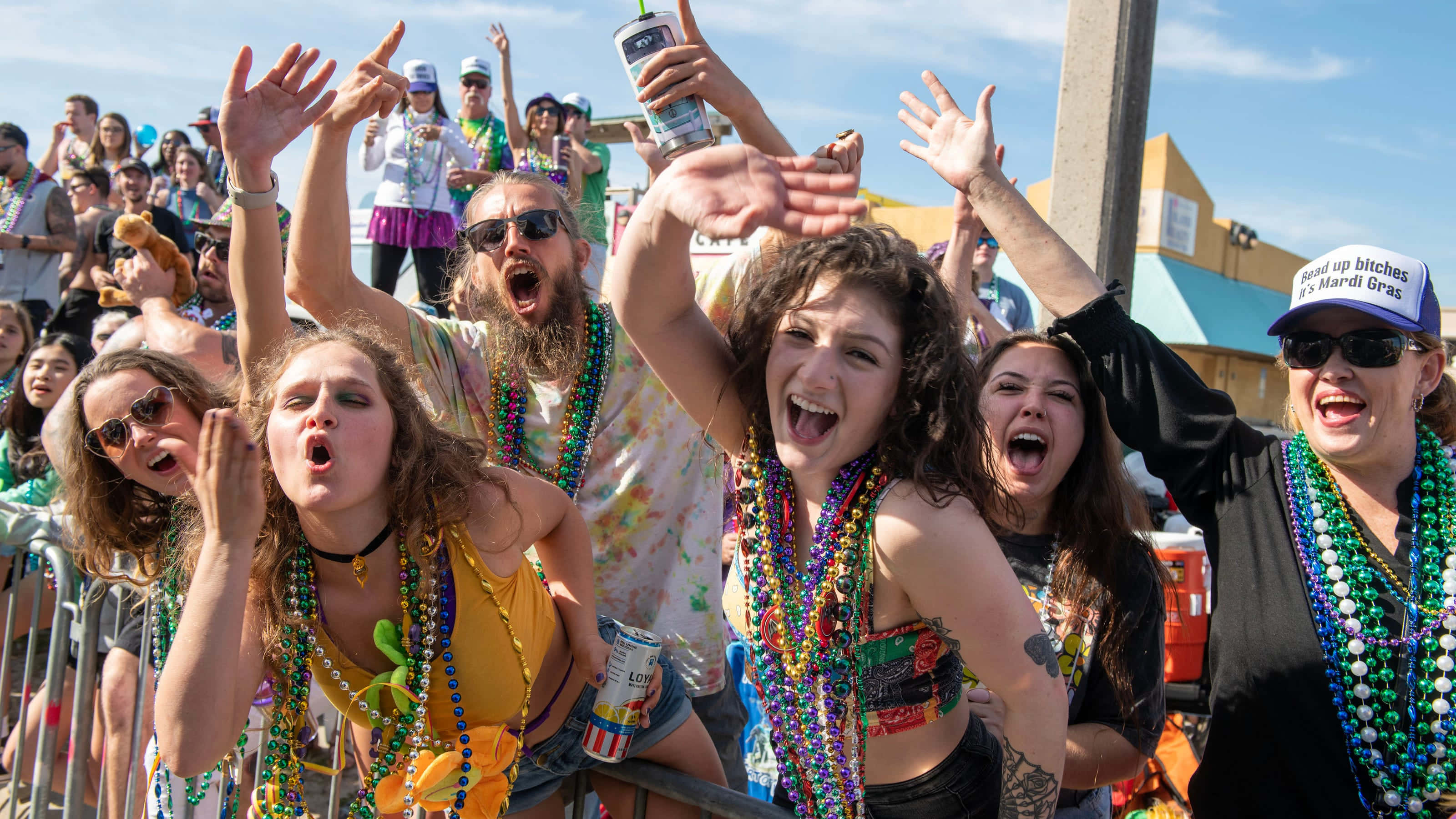 Mardi Gras 桌布 壁紙 — 在狂歡節上，充滿活力的人群在晴朗的天空下歡呼，手持彩色珠子和飲料。