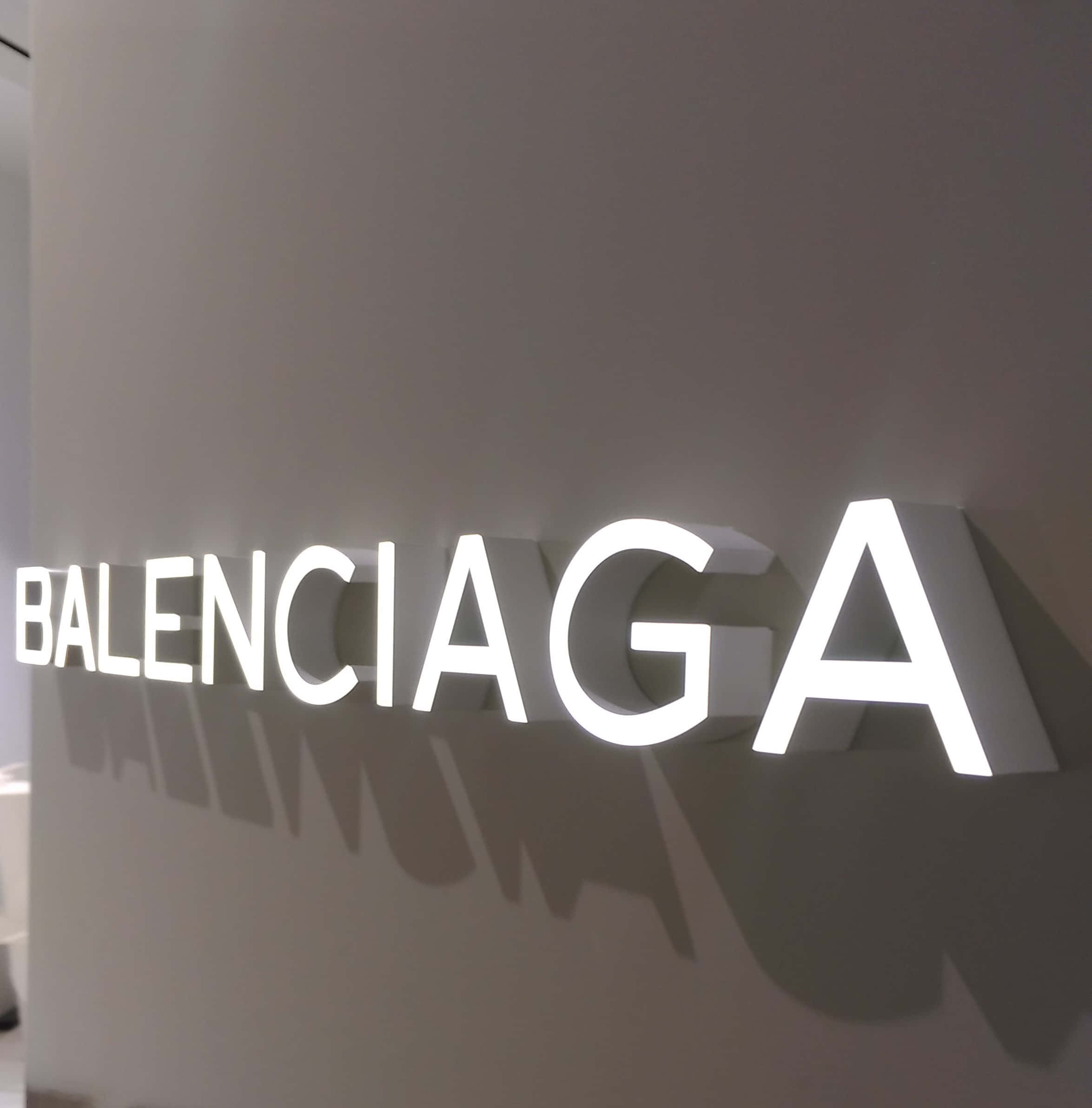 Balenciaga 桌布 壁紙 — 在現代零售空間中，一個極簡風格的白色發光 Balenciaga 標誌在啞光灰色牆壁上投下銳利的陰影。