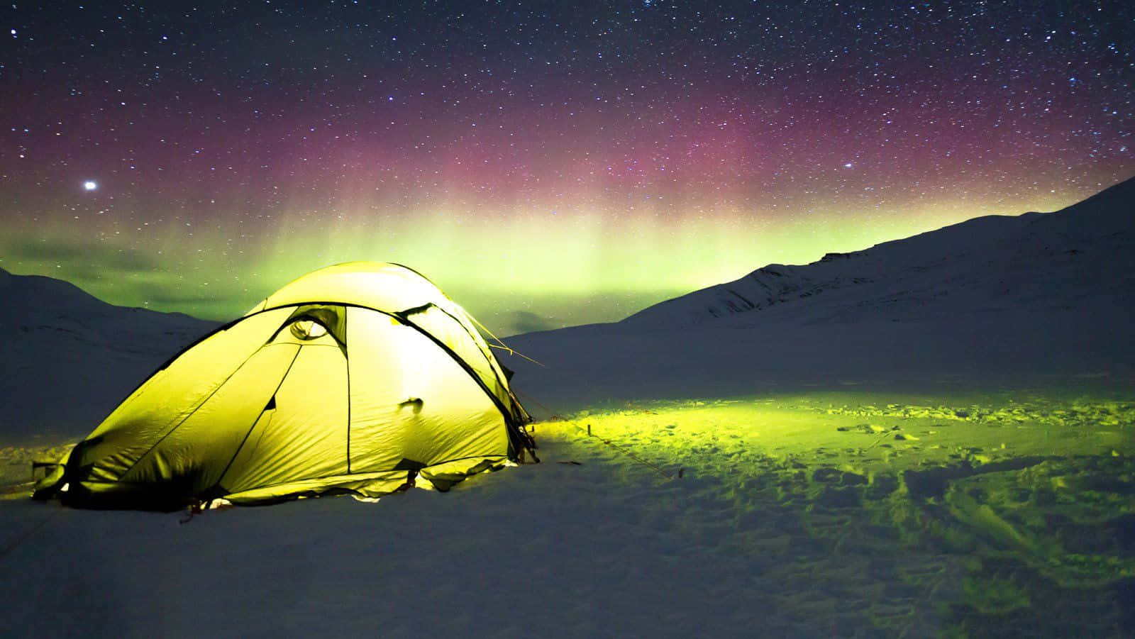 Winter Camping 桌布 壁紙 — 在白雪覆蓋的山巒中，一盞發光的黃色帳篷，上方是橫跨星空的鮮綠色和紫色極光。