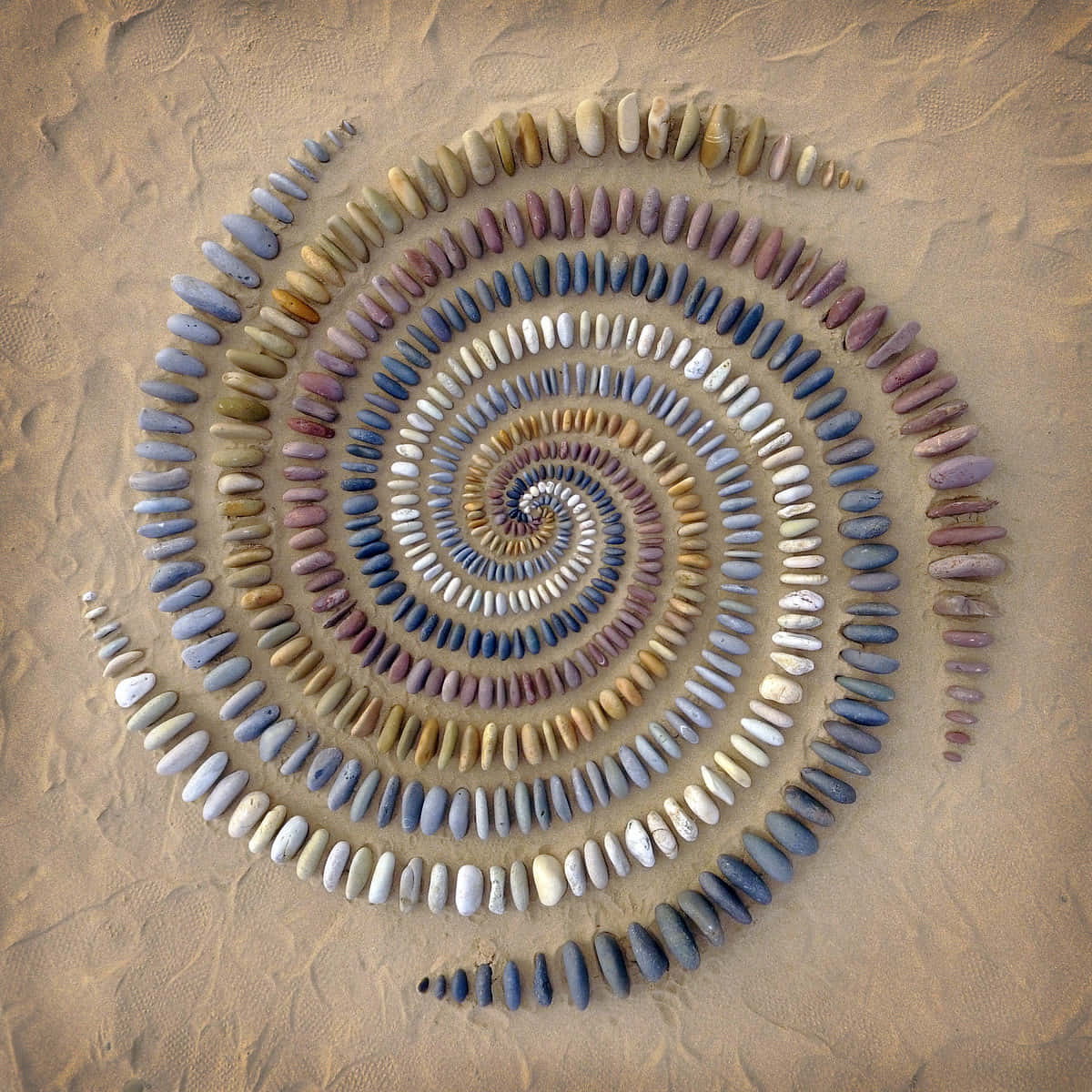 Pebble Art 桌布 壁紙 — 在紋理沙地上排列的光滑多色石頭形成的迷人螺旋，創造出平靜的幾何圖案。