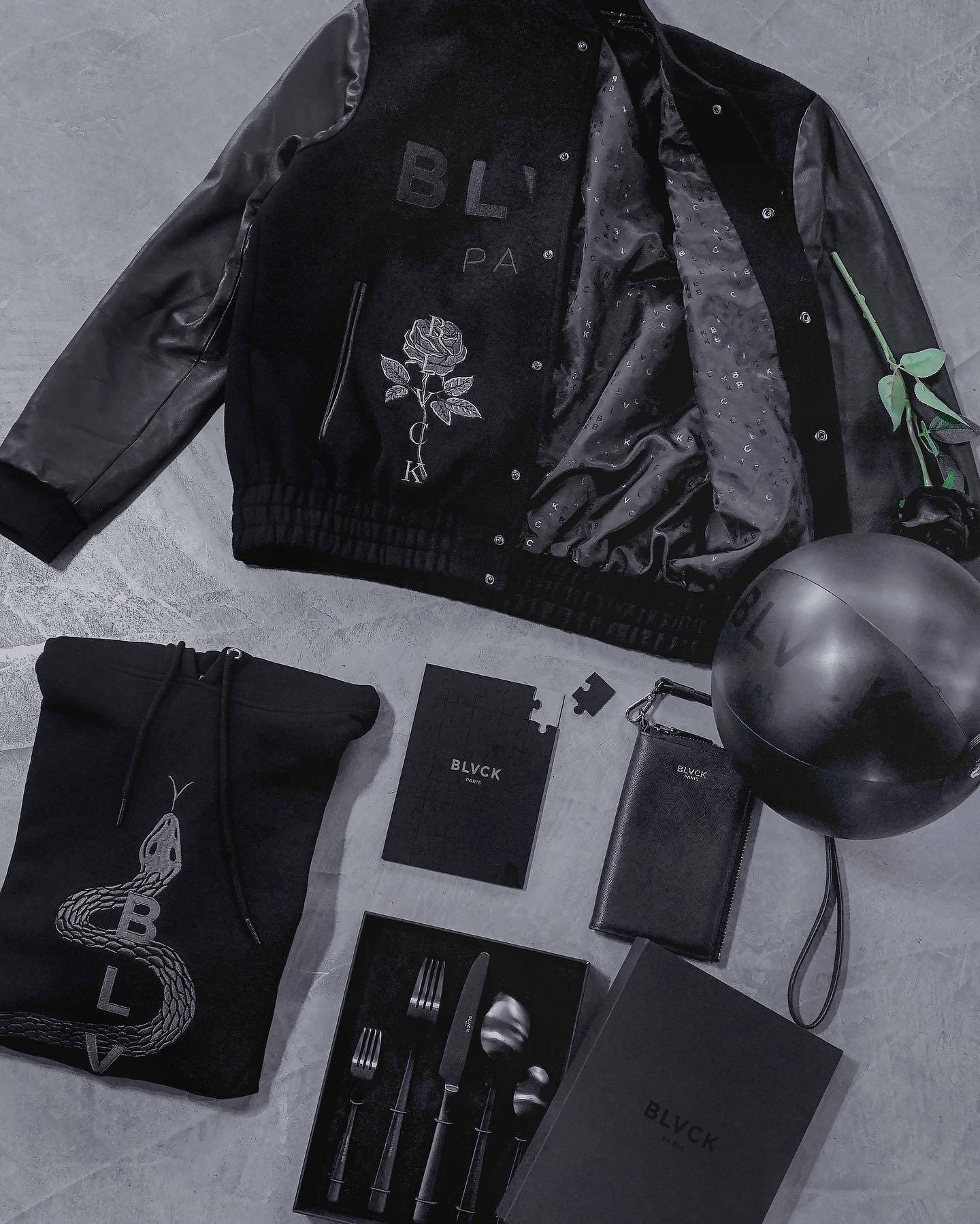 Blvck Paris 桌布 壁紙 — 在紋理灰色表面上，一件帶有玫瑰刺繡的黑色轟炸機外套，周圍環繞著相配的品牌配件，形成單色平鋪佈局。