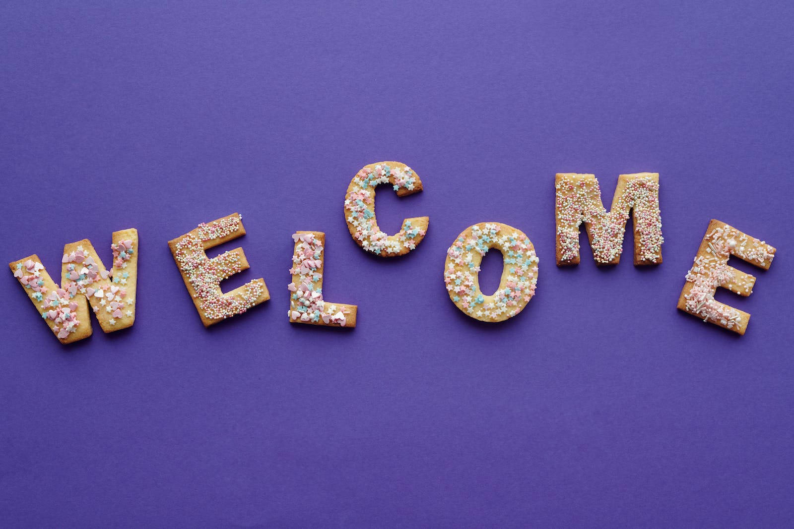 Welcome 桌布 壁紙 — 在純紫色背景上，用色彩繽紛的糖霜曲奇拼出「WELCOME」一詞。