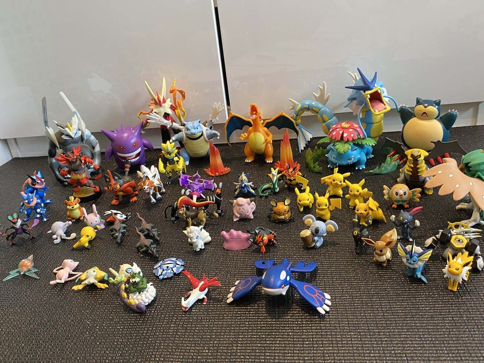 Pokemon Figures 桌布 壁紙 — 在素色牆壁前，暗色紋理表面上排列著色彩繽紛的寶可夢公仔收藏。
