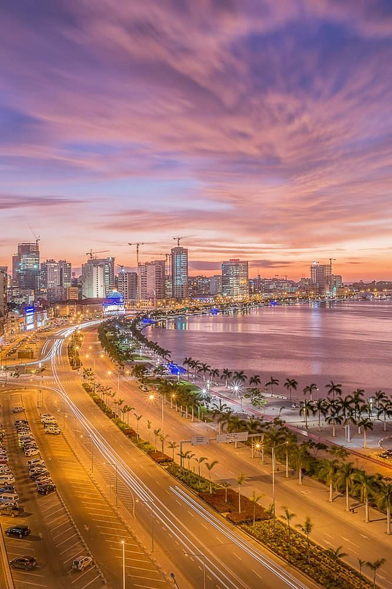 Angola 桌布 壁紙 — 在紫色和橙色天空下，海濱道路上車燈流光溢彩的活力城市景觀。