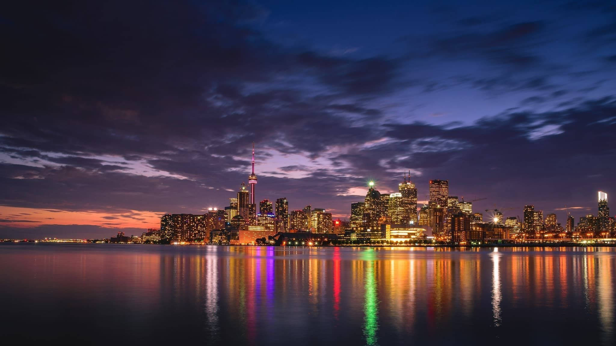 Ontario 桌布 壁紙 — 在紫色和橙色天空下，色彩繽紛的燈光倒映在平靜水面上，充滿活力的城市天際線在黃昏時分。